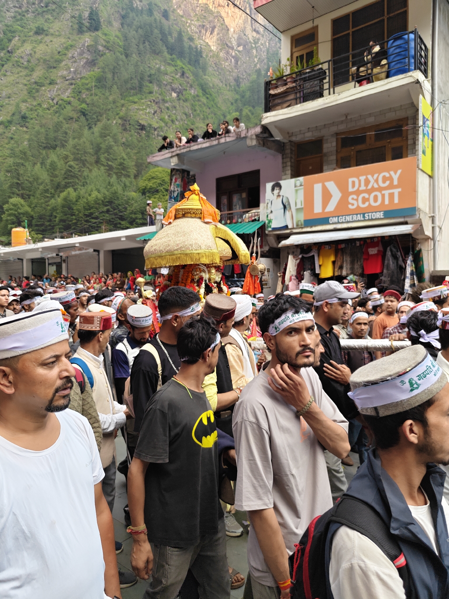 The image captures a lively religious procession unfolding on a crowded street in Manikaran, India, set against a backdrop of steep, forested mountains. Numerous people, primarily men, are either participating in or observing the event under an overcast sky.
At the center of the procession is an ornate deity palanquin, richly adorned with gold-colored, embroidered fabric and topped with a gilded spire, being carried through the throng. Many of the participants wear traditional Himachali caps, while a significant number also sport white headbands. These headbands prominently display text that appears to say "ॐ गड़पती श्री ईश्वर" (Om Gadpati Shri Ishwar) along with an Om symbol and a green emblem, possibly a leaf or tree.
The street is lined with multi-story buildings, some with balconies where onlookers gather to view the procession. One prominent orange sign advertises "DIXCY SCOTT OM GENERAL STORE" and features an image of a man. Other clothes are visible for sale in the shop. The scene suggests a community-wide religious festival or celebration, characterized by a sense of devotion and collective participation in the mountainous setting. The natural light indicates it is daytime, possibly morning or afternoon, with diffused lighting due to the cloud cover.