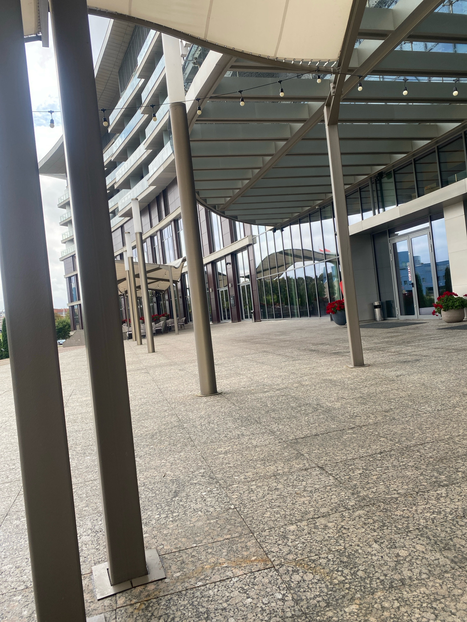 A modern building with a glass facade is visible from the exterior. The building has multiple floors with balconies and is set against a cloudy sky. A covered walkway with a fabric roof extends from the building, supported by metal columns. String lights hang from the structure. The ground is paved with speckled granite tiles. In front of the building, there are large planters with red flowers. A few tables and chairs are visible under the covered walkway, suggesting an outdoor seating area. The scene appears to be a commercial or residential complex. The location is not specified in Minsk, Belarus.