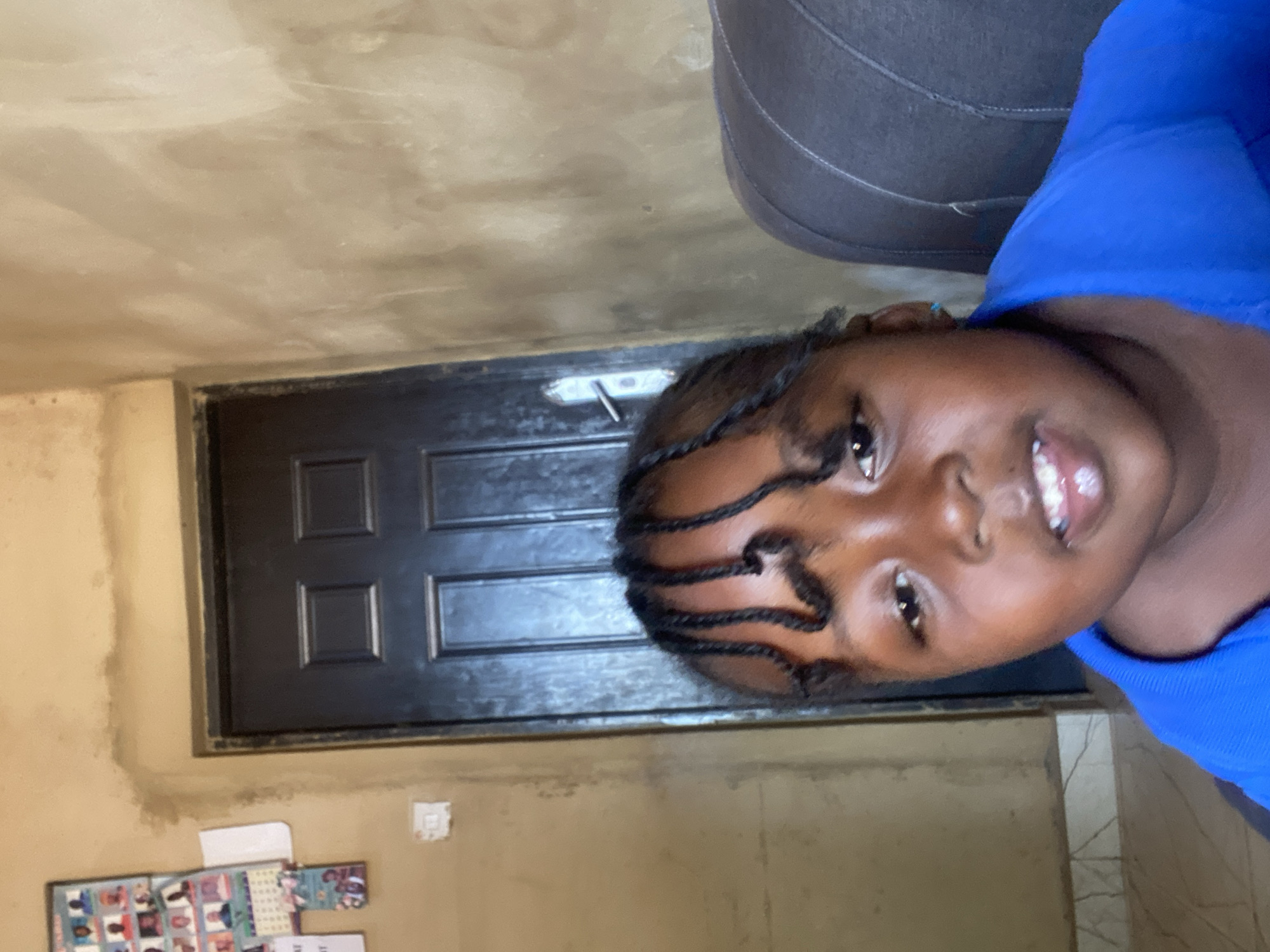An upward-angled, candid portrait captures a young person, likely a girl, smiling brightly indoors in Kuru Dungus, Nigeria. The subject, positioned centrally and looking directly at the camera, has dark skin, expressive dark eyes, and a broad smile revealing her teeth. Her hair is styled in neat braids, with some strands framing her forehead. She wears a vibrant blue ribbed top, and a small, possibly blue, earring is visible in her right ear.
The background reveals an interior space with textured, light brown walls, suggesting an unpainted or rustic finish. Behind the subject, a dark brown or black panel door with multiple recessed squares is prominently featured, indicating a room or entrance. A silver-colored door handle or lock mechanism is visible on the door. To the upper right, a section of a dark grey, possibly upholstered, piece of furniture like a sofa or chair is partially visible.
In the lower left, a wall calendar or poster with numerous small portrait photographs and some text is affixed to the wall. Faintly visible text includes "PEOPLE". Nearby, several white papers or notes are also attached. A white electrical outlet or switch is visible on the wall to the right of the calendar. The floor, partially seen in the lower right, appears to be made of light-colored, possibly veined, tiles. The overall impression is a casual, everyday scene, with the person appearing happy and relaxed.