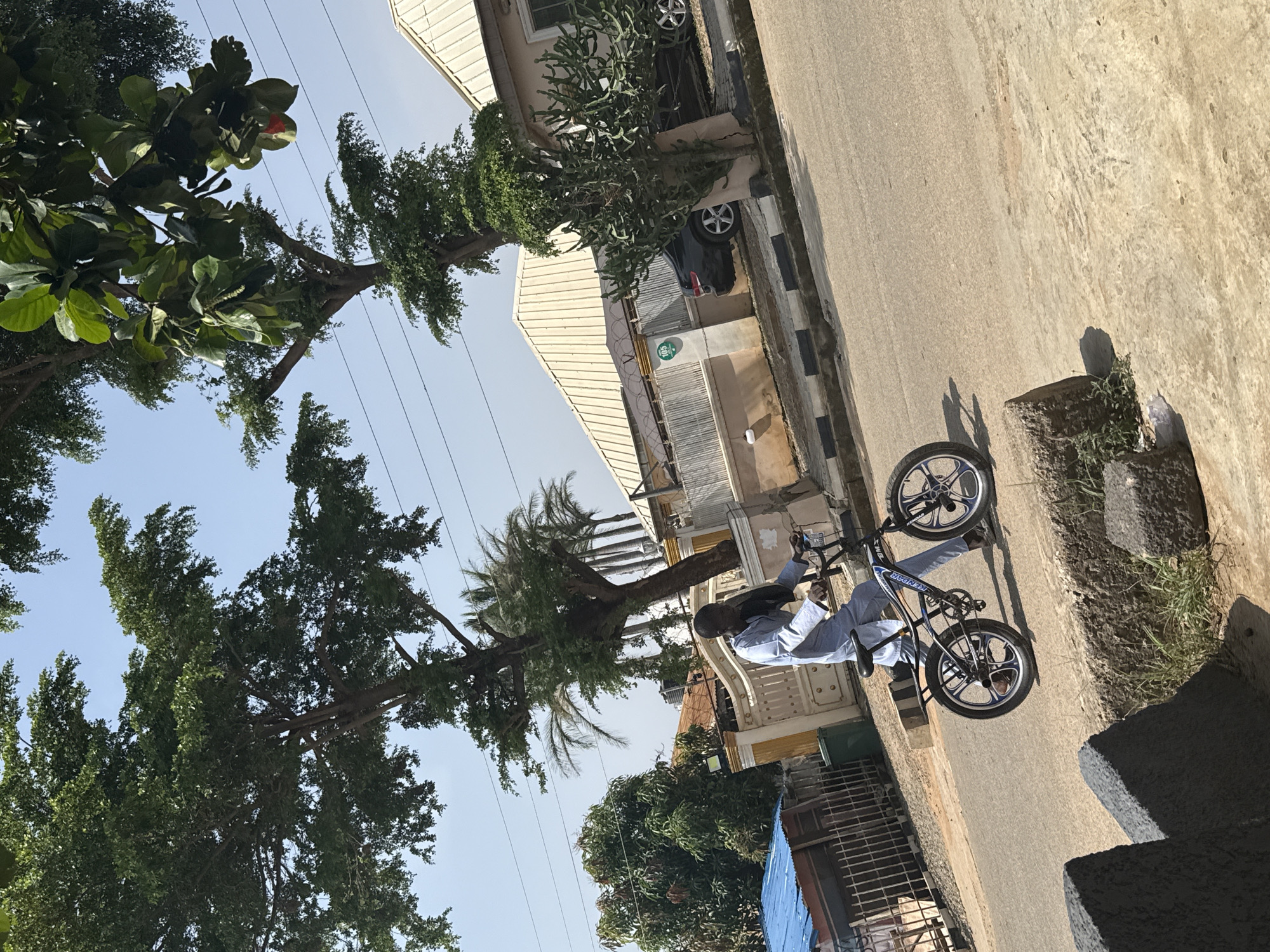 A person is riding a BMX bicycle on a paved street in what appears to be a residential area. The person is wearing a light blue long-sleeved shirt and light blue pants. The bicycle is black with silver rims.
The scene is outdoors with a clear blue sky. Lush green trees with large leaves are prominent in the foreground and frame the image, some with branches reaching towards the sky. In the background, there are buildings with light-colored walls and corrugated metal roofs. Some of these buildings have windows and balconies. A dark-colored car is parked near one of the buildings.
The street is made of concrete and has some wear and tear. A curb with black and white markings separates the street from a concrete walkway. Several electrical wires are visible overhead, strung between poles.
The lighting suggests it is daytime, likely mid-morning or afternoon, with bright sunlight casting shadows. The overall mood is casual and relaxed. The location context of Abuja, Nigeria is suggested by the architecture and vegetation. No visible text is discernible in the image.