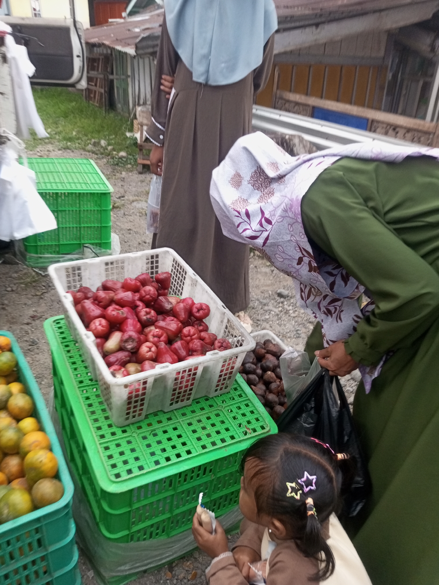 A street market scene in Pandansari, Indonesia, features an outdoor display of produce. In the foreground, a young child with black hair styled in pigtails adorned with star-shaped clips is partially visible. The child is looking to the left and holding a small item in their right hand. To the child's right, a woman in a green abaya and a patterned headscarf is bent over, holding a black plastic bag. Behind her, a white crate overflows with bright red fruits, possibly rose apples. Adjacent to this, another crate contains dark, oval fruits, likely salak. Further to the left, a green plastic crate holds a pile of oranges and other citrus fruits. In the background, a person in a brown abaya and a light blue headscarf stands near a white vehicle with its door open. The ground is covered in gravel and small stones. The overall environment suggests a warm climate.