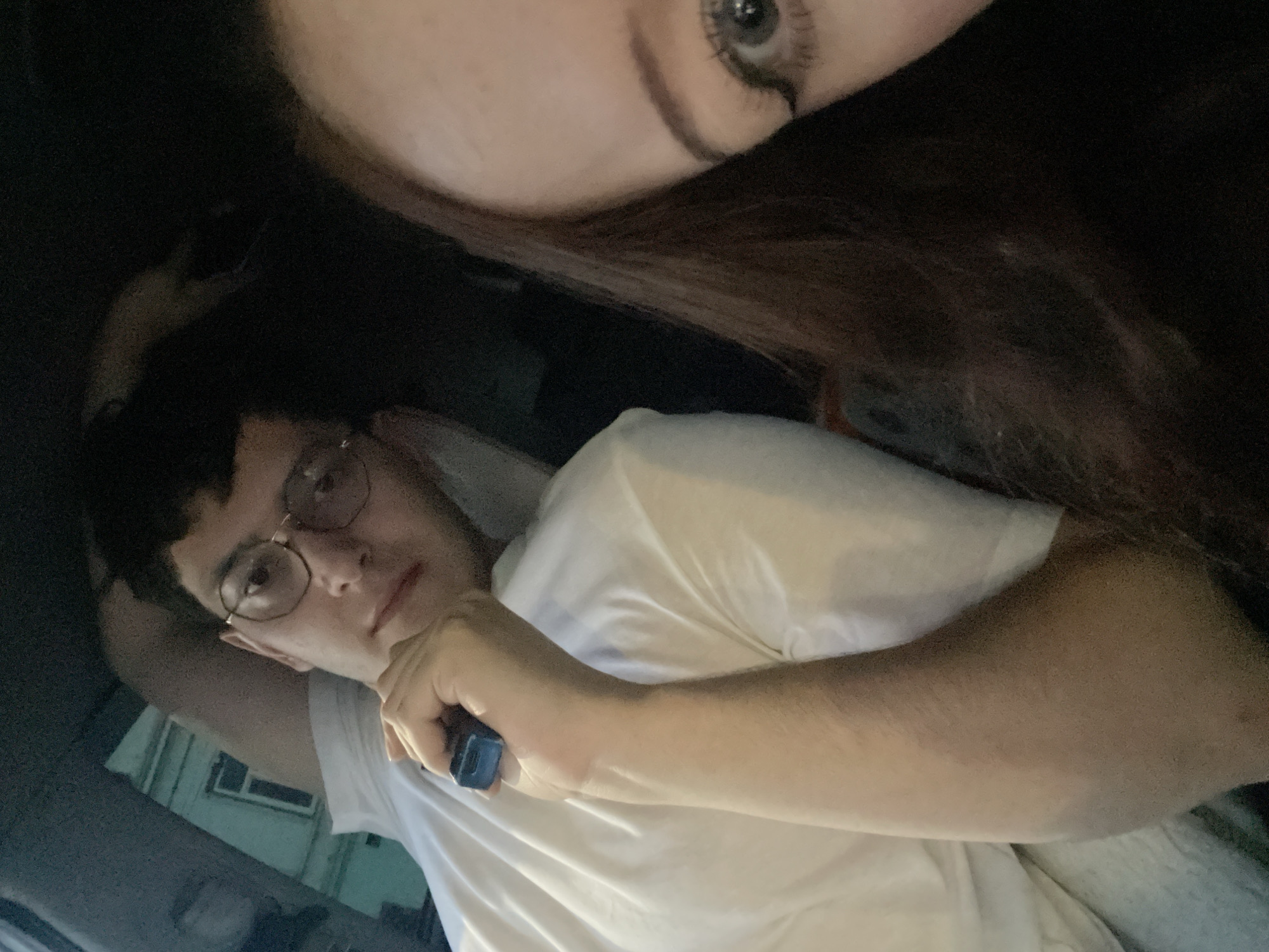 This is a tilted, close-up shot featuring two individuals in a dimly lit setting. In the foreground, a young man with dark, short hair and thin-framed glasses looks directly at the camera with a neutral to slightly thoughtful expression. He is wearing a plain white t-shirt. His right hand holds a small, rectangular, dark blue device, and his left arm is bent, with his hand seemingly behind his head or neck.
Above him, partially cropped in the upper right corner of the frame, is a portion of a woman's face, primarily her eye (which appears light-colored, possibly blue or gray, with visible eyelashes) and forehead, surrounded by dark hair. Her proximity suggests she is either taking the photo or leaning very close to the camera.
The background behind the man is dark and indistinct, suggesting an indoor environment or a vehicle interior at night. A faint, light-colored architectural detail, possibly a window frame or part of a building, can be vaguely discerned to the left of the man's head, illuminated by an unseen light source. The overall lighting is low, creating a soft and intimate atmosphere. No text is visible in the image.