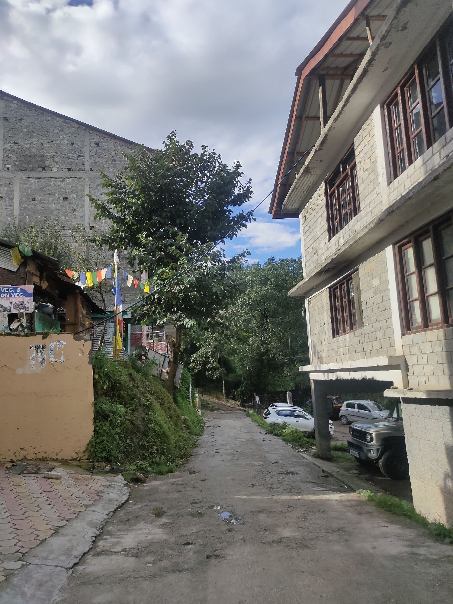The image depicts a narrow, unpaved road in Manali, India, flanked by buildings and vegetation. On the right, a multi-story building with a grey brick facade and wooden-framed windows is prominently featured. Its roof is covered with corrugated metal. In front of this building, two cars are parked: a light-colored SUV on the left and a darker SUV on the right. A small three-wheeled auto-rickshaw is also visible behind the cars.
On the left side of the road, a smaller, older structure with a yellow-painted wall stands. Prayer flags in various colors (blue, white, yellow, red, green) are strung across the area in front of this structure. Above this, a large, grey stone building with a sloping roof can be seen. Lush green trees and vegetation cover the hillside between the road and the buildings. The sky is partly cloudy with a mix of blue sky and white clouds. The road surface is uneven and appears to have some puddles. The overall impression is of a residential or commercial area in a hilly region.