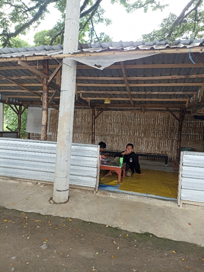 The image depicts a rustic, open-sided structure in Kepung, Indonesia, likely serving as a shelter or a small roadside eatery. The structure is built with a framework of wooden beams and posts, a corrugated metal roof, and walls primarily made of woven bamboo. The front-left section features a low barrier made of corrugated metal sheeting. A prominent, thick concrete or stone pillar stands in the foreground, appearing to be a central support for the roof.
Inside the shaded area, two individuals, possibly young males, are relaxing. They are seated on a yellow mat spread across the floor. The person on the right, wearing a black long-sleeved shirt with some white detailing, is looking towards the camera with a relaxed expression, holding a green plastic cup with a straw. He appears to be kneeling or squatting. To his left, another person is partially visible, seated at a low, small wooden table. A white panel or board is visible on the interior left wall.
The setting suggests a daytime scene, with soft, diffused natural light indicating either an overcast sky or significant shade from the surrounding foliage. Trees and lush greenery are visible in the background above the structure. The ground outside is dirt, scattered with leaves and small rocks. The scene conveys a sense of a casual, informal outdoor resting spot. No discernable text is visible within the image.