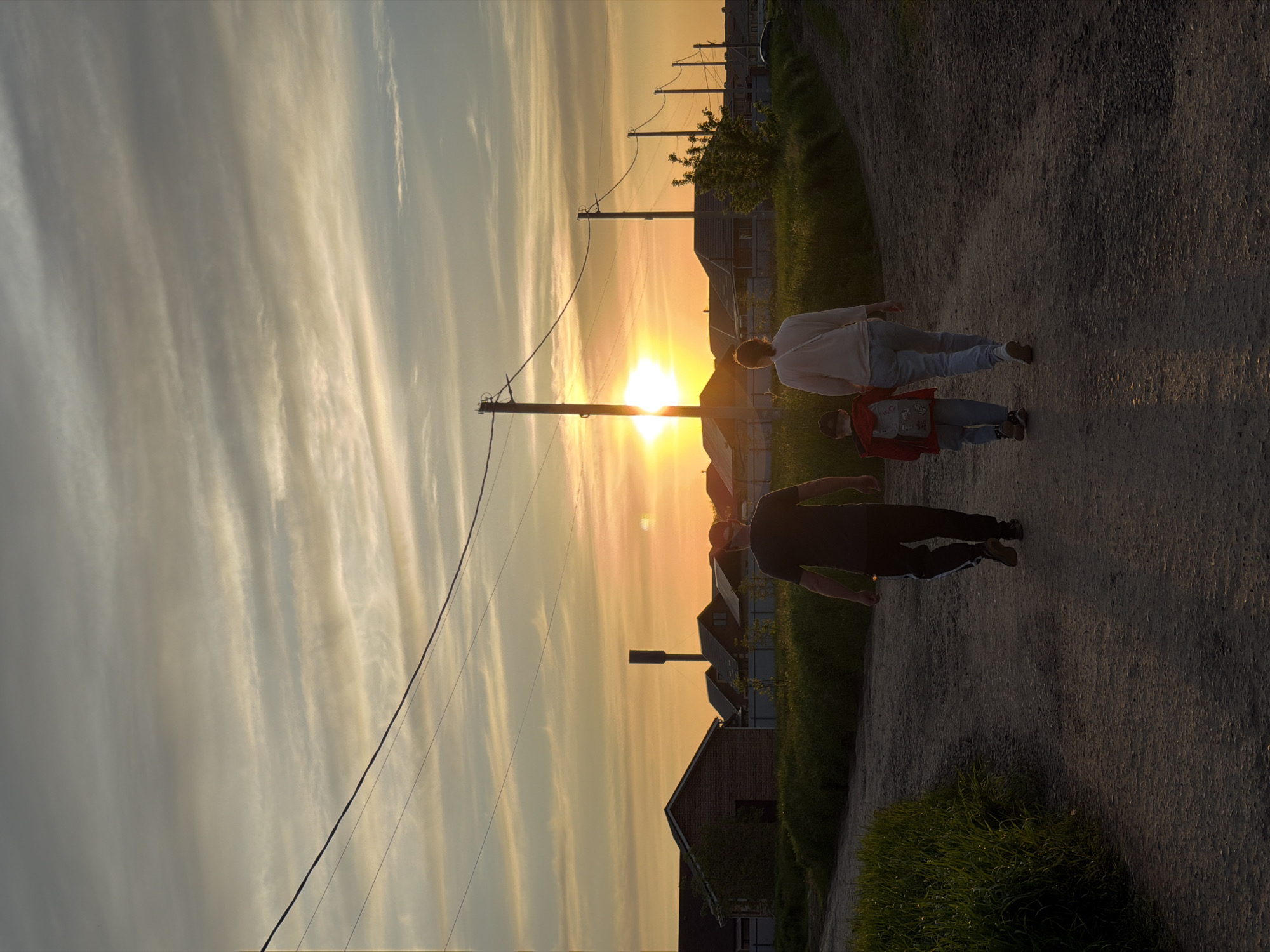 The image depicts a sunset in Rossiyskiy, Russia. Three people are walking on a dirt road alongside grass and utility poles. Houses are visible in the background, silhouetted by the sun. The sky is filled with light clouds illuminated by the setting sun.