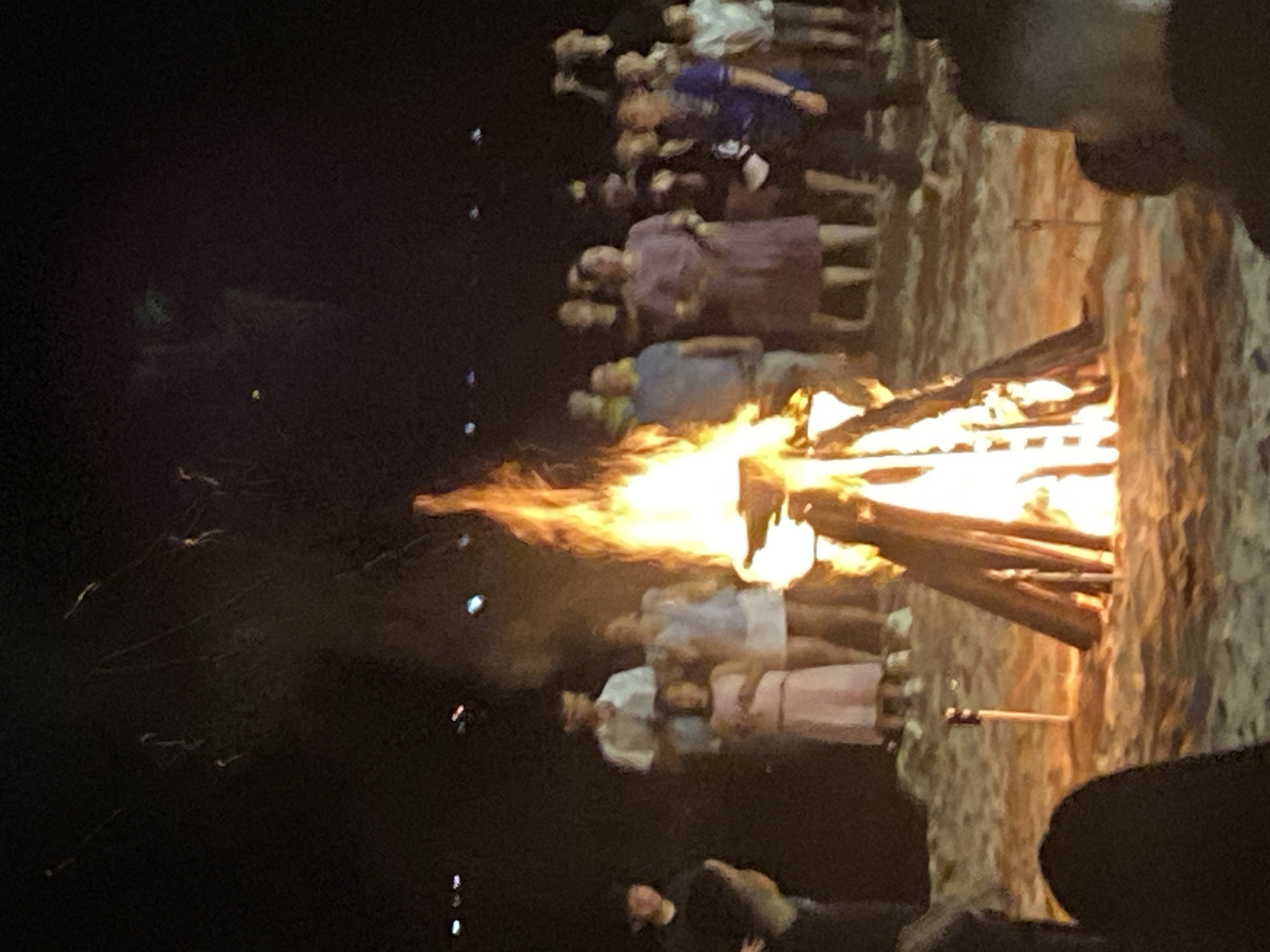 A group of people are gathered around a bonfire on a sandy beach at night. The bonfire is large and is made of stacked logs. Flames are leaping from the top of the bonfire. In the background, there are lights from a city or town. The people are of various ages and are dressed in casual clothing. Some of them are looking at the bonfire, while others are talking to each other. The scene appears to be a festive gathering. The location is identified as Phu Thuy Ward, Vietnam.