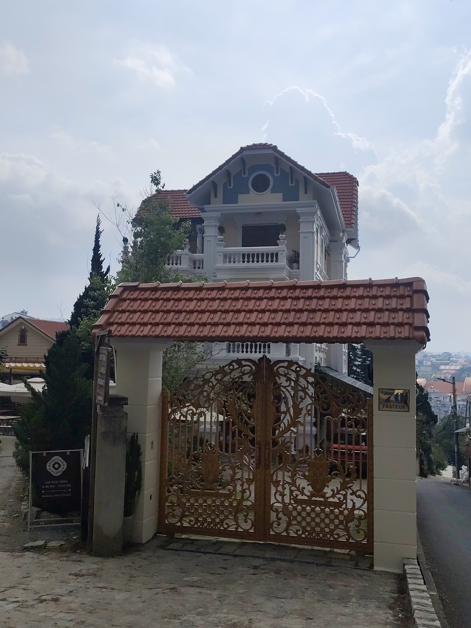 The image shows the entrance to a property located on Pasteur Street in Da Lat, Vietnam. A decorative, golden gate stands between two white pillars, covered by a tiled roof structure. To the left of the gate is a sign with Vietnamese script, partially obscured. The house behind the gate is a two-story building with a red tile roof and a balcony. The sky is overcast. In the distance, buildings can be seen, indicating an urban area. A road slopes downward to the right of the gate.