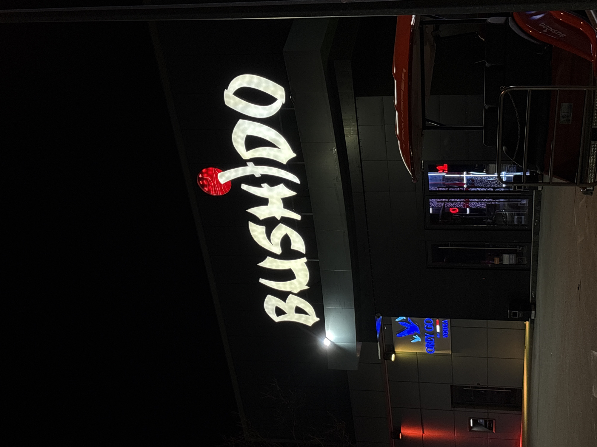 At night, an illuminated sign for "BUSHIDO" is prominently displayed vertically on a dark building in Plovdiv, Bulgaria. The letters are bright white, except for a red circle at the top, perhaps as part of the logo. Below the sign is a Grey Goose Vodka sign lit with blue and white. The building appears modern with dark windows. A red vehicle is partially visible above the Bushido sign. The ground is concrete.