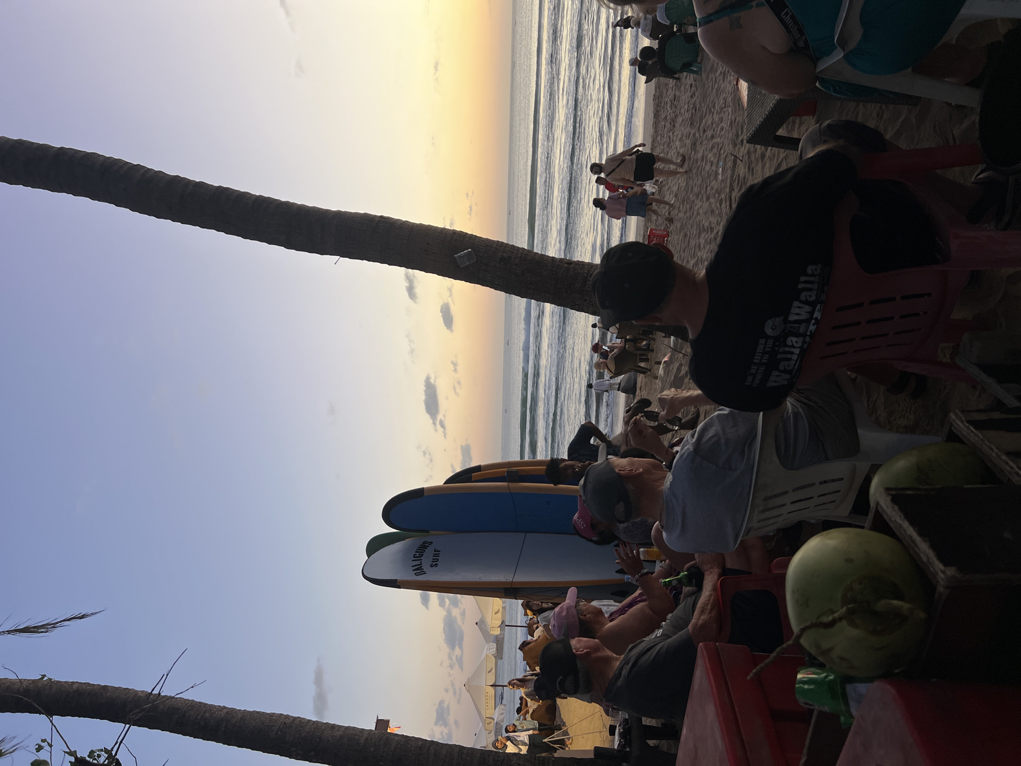 A group of people are seated at tables on a sandy beach in Kuta, Indonesia, watching the sunset over the ocean. Several surfboards are propped up on the beach. Palm trees frame the scene. The sky is a gradient of orange, yellow, and blue, with scattered clouds. In the foreground, a few people are visible in close-up, facing away from the camera, with drinks on their table. The overall atmosphere is relaxed and leisurely, typical of a beachside gathering.