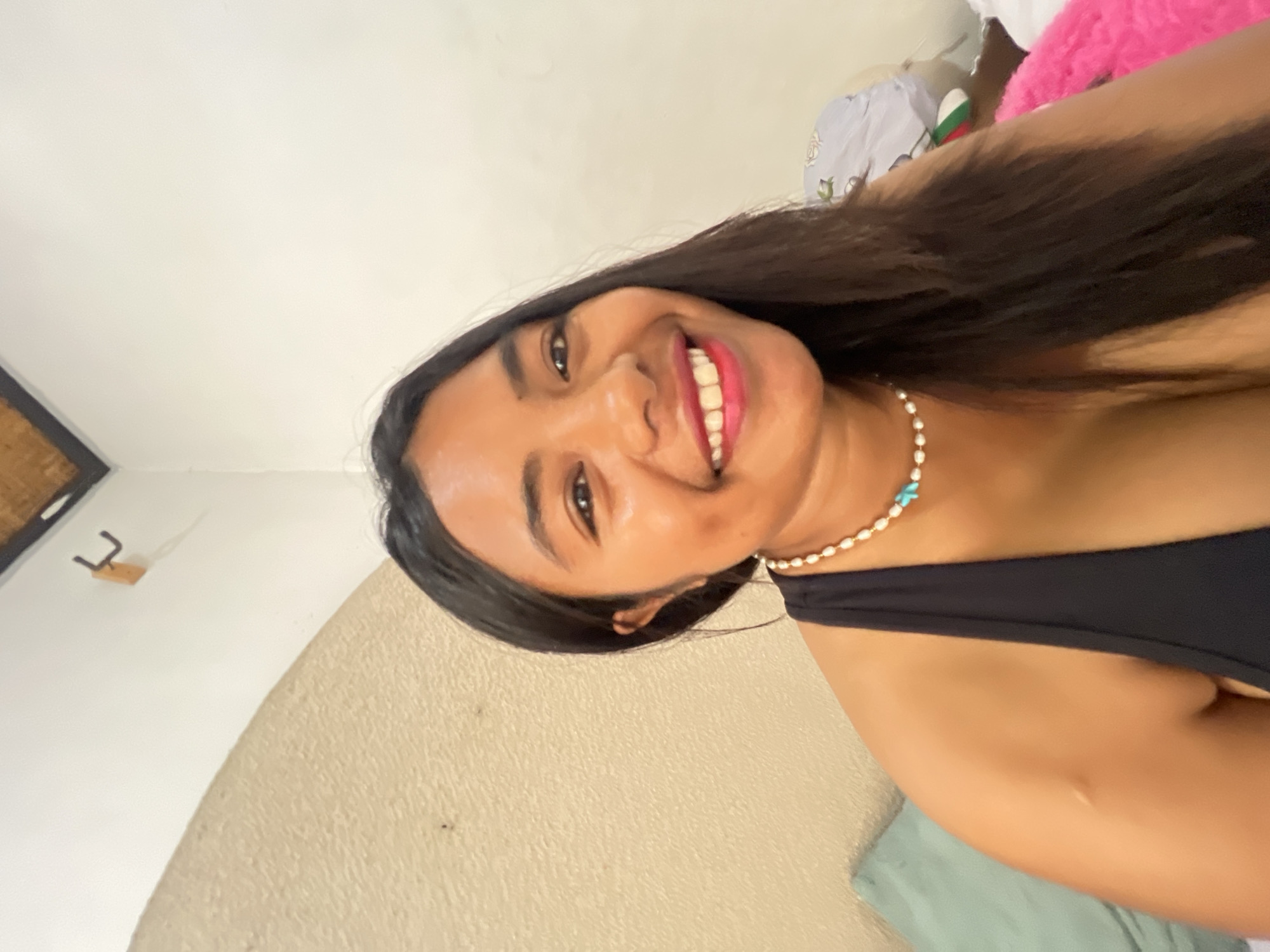 A young woman with dark hair and a bright smile is captured in a selfie taken indoors. She is wearing a black tank top and a pearl necklace with turquoise accents. The background suggests a bedroom with a textured wall, a ceiling with a bamboo pattern, and some bedding. A hook is visible on the wall. The lighting is natural and illuminates her face, highlighting her rosy cheeks and full lips.
