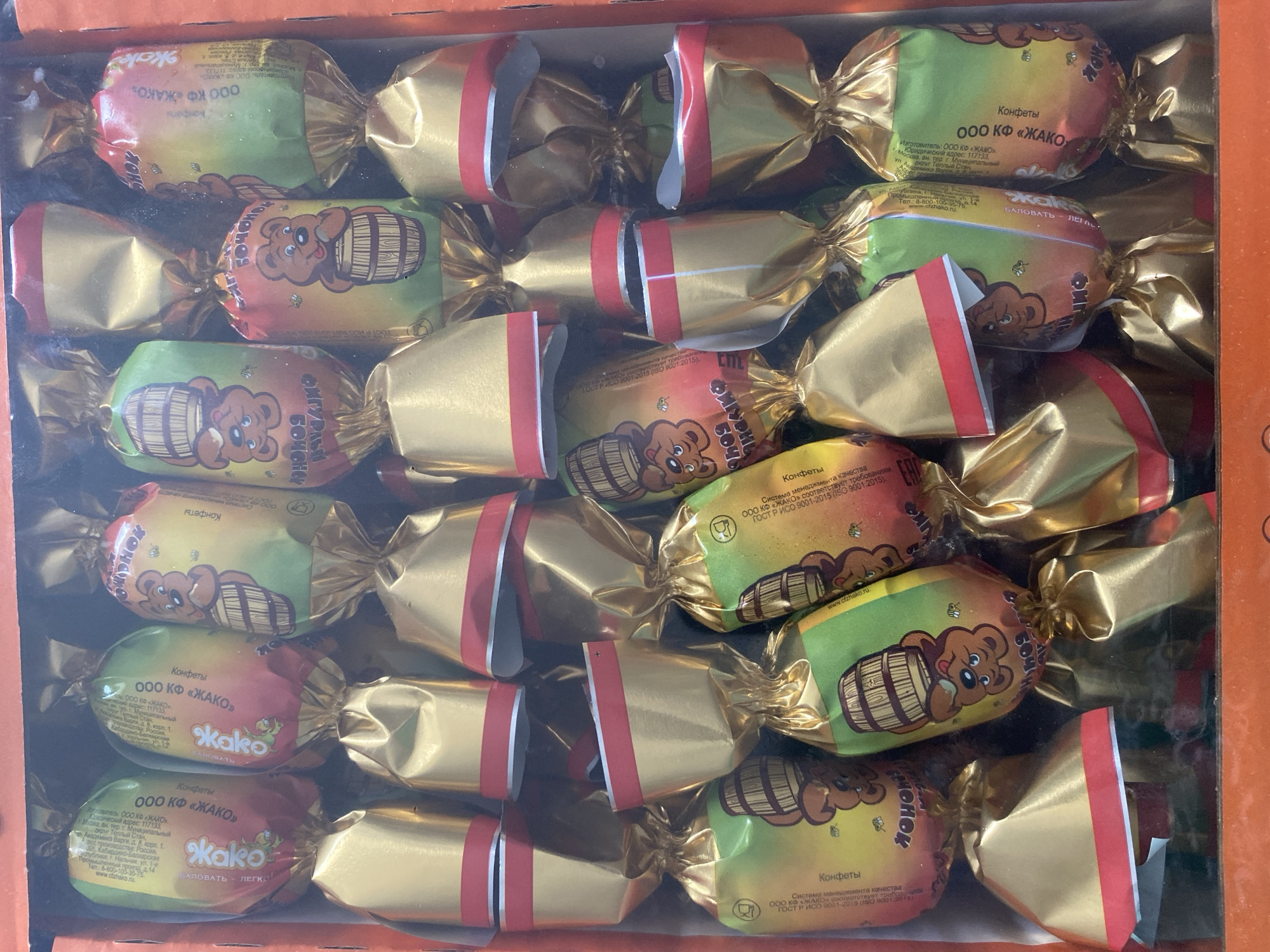 A box of individually wrapped candies is shown, with the candies arranged in rows. The wrappers are predominantly gold with red accents, and some feature colorful illustrations of cartoon bears holding barrels and bees. Text on the wrappers is in Cyrillic. The box is orange. The image does not provide any information about the city of Chontaul, Russia, or any specific location within it.