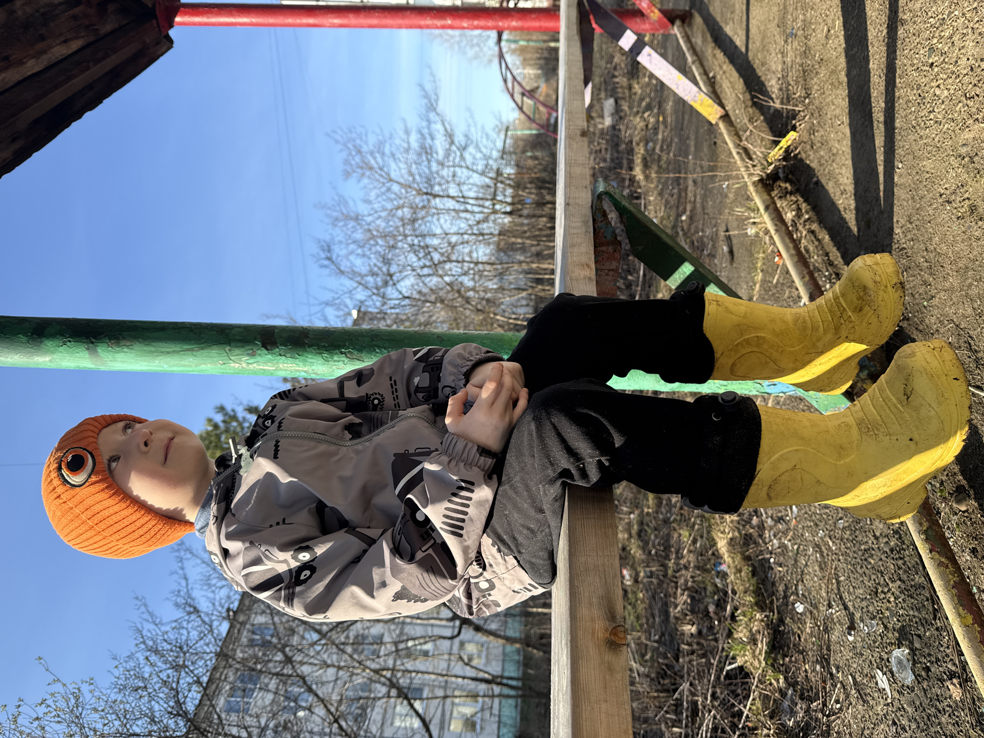 A child wearing an orange beanie, a patterned gray jacket, black pants, and yellow rubber boots is sitting on a wooden beam. The beam is part of a playground structure with green and red bars. The background includes leafless trees, a blue sky, and a building. The location is likely a playground in the city of Chaikovsky, Russia.