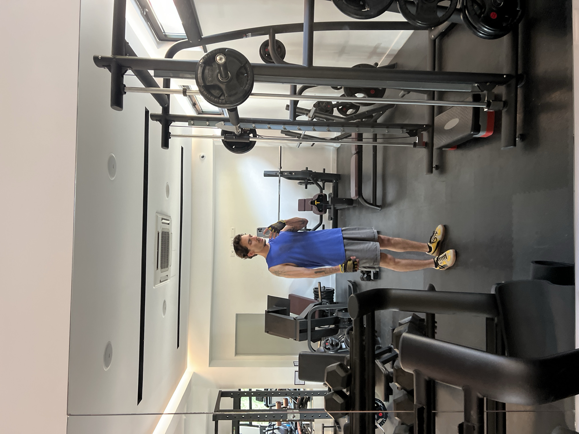 The image depicts a man in a private gym, likely in a luxury home in Boreti, Montenegro. The gym is well-equipped with various weightlifting machines, including a squat rack with weights, a bench press, and other fitness equipment. The man is wearing a blue t-shirt, gray shorts, yellow shoes, and workout gloves. He is holding a smartphone and appears to be taking a selfie or video. The gym is reflected in a large mirror, showing the layout of the room and the equipment. The room is well-lit with a modern design, including recessed lighting and a skylight.