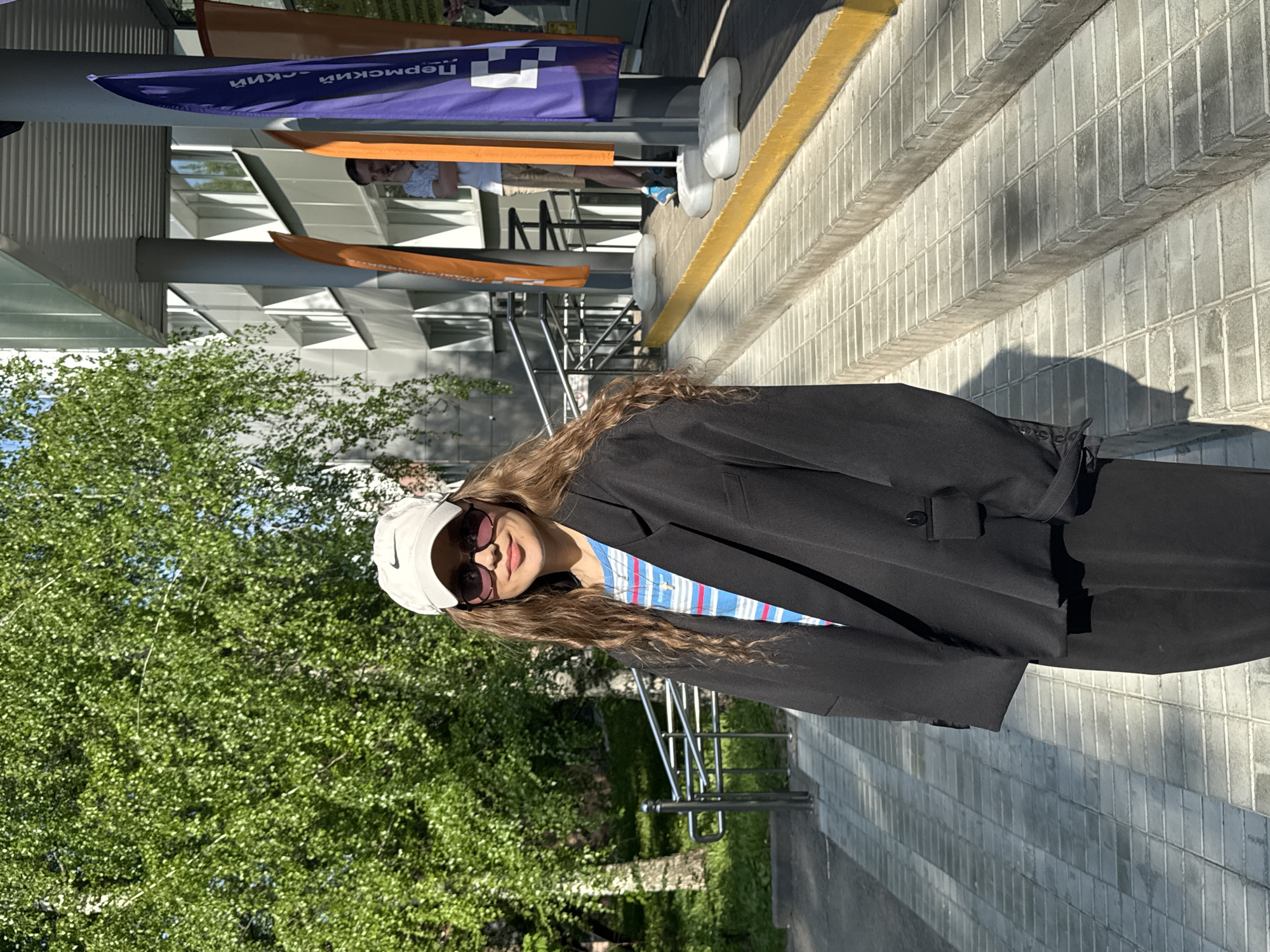 A woman with long, wavy brown hair, wearing a white baseball cap, sunglasses, a striped shirt, and a black blazer, stands on a sidewalk in Perm, Russia. She is facing the camera and smiling. In the background, there is a modern building, green trees, and signage in Russian ("Пермский"). Other people are visible in the background. The sidewalk is made of light-colored brick, and there are metal railings along the side.