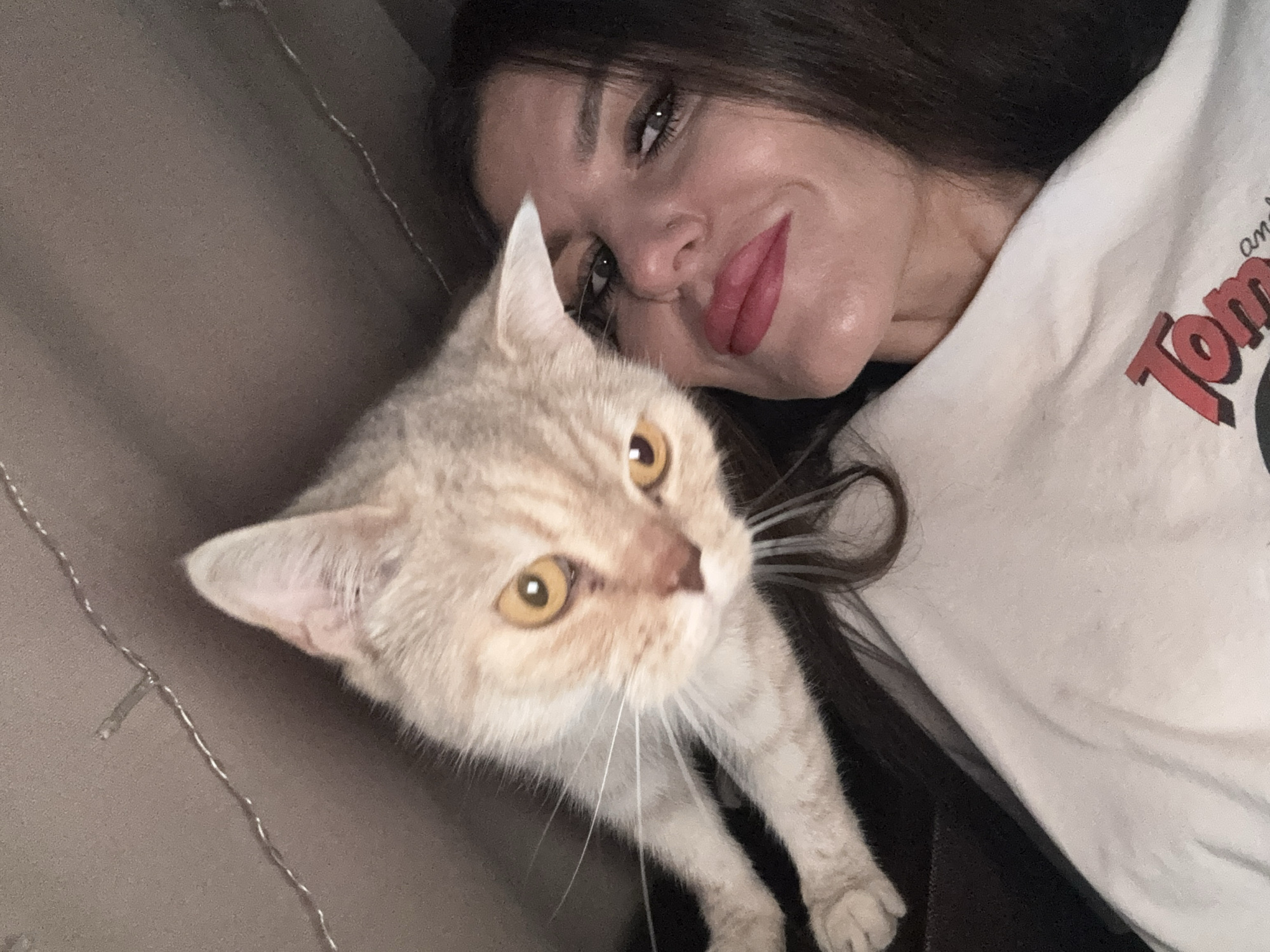 This is a close-up, low-angle selfie of a woman and a cat. The woman, with dark hair and bold pink lipstick, is looking towards the camera with a slight smile. The cat, with light orange fur and yellow eyes, is positioned in the foreground, its face very close to the camera, appearing to be interacting with the woman. The background is dark and out of focus, with some faint string lights visible near the top of the frame. The setting appears to be indoors, possibly a home environment. There is no discernible connection to the city of Chaikovsky, Russia.