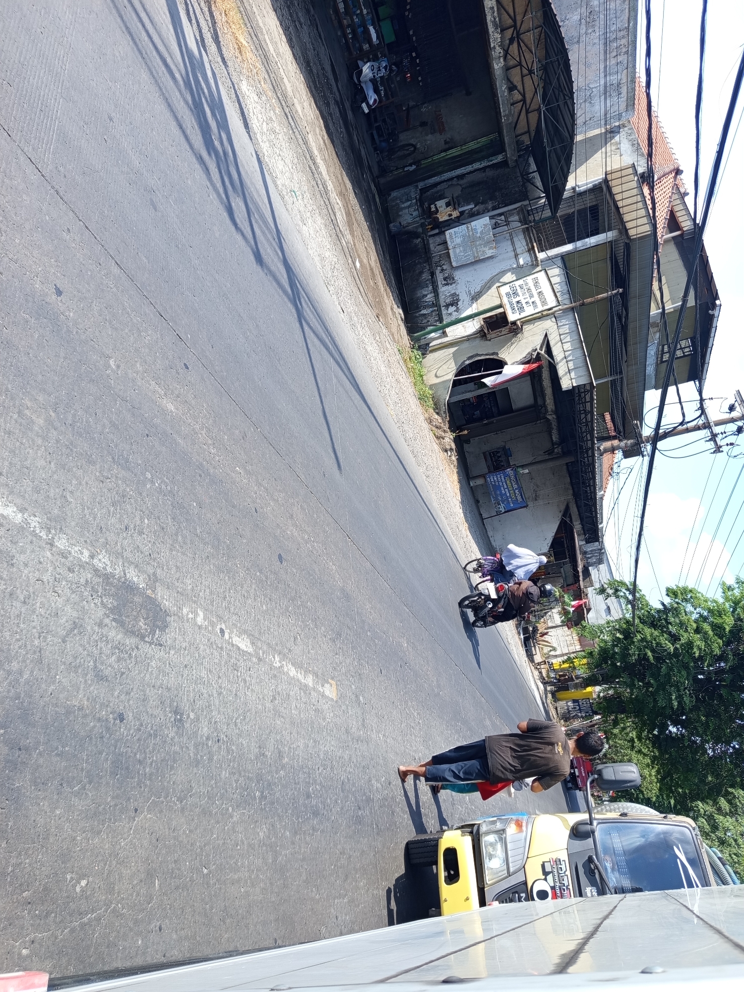 The image, captured in Sidomulyo, Indonesia, depicts a street scene on a sunny day. The foreground is dominated by the grey asphalt of a road, marked with a white dashed line. In the mid-ground, a man is walking barefoot on the side of the road, wearing a dark t-shirt and dark pants. He is walking towards a yellow truck with a reflective windshield, positioned on the right side of the road. The truck has visible signage on its windshield. Further down the road, two motorcycles are visible: one in front of the other, each carrying passengers. The motorcycle in the foreground has a rider and a passenger wearing a white headscarf. The background features a two-story building with a covered walkway and an awning. Various signs, some with red and white Indonesian flags, are attached to the building. Electrical wires are visible above the building, and a dense green tree is situated to the right of the building. The scene is oriented with the road leading away from the viewer at an angle.