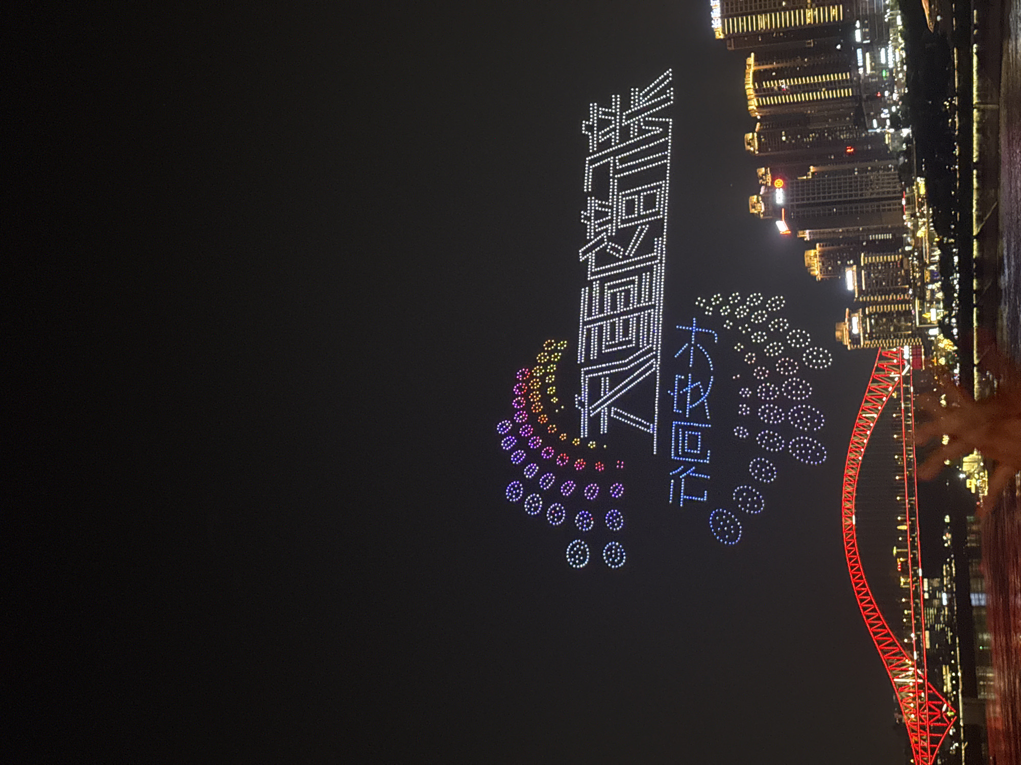 An aerial nighttime drone show illuminates the sky above Yuzhong District, China, with a vibrant display of lights. A large, multi-colored arc of illuminated circles is positioned in the upper left of the frame, transitioning from white to purple, pink, orange, and yellow. To the right of the arc, large white characters are formed by a dense formation of lights, appearing as illuminated text in the sky. Below this text, a cluster of blue, circular light formations is visible. In the lower portion of the image, the Chongqing Yangtze River Bridge is prominently featured, illuminated in red. The bridge spans a dark body of water, likely the Yangtze River, which reflects some of the city's lights. The background consists of a cityscape with numerous tall buildings, many of which are lit with golden and yellow lights. The overall scene depicts a festive night with a drone light show as the focal point, set against the backdrop of a modern Chinese city.