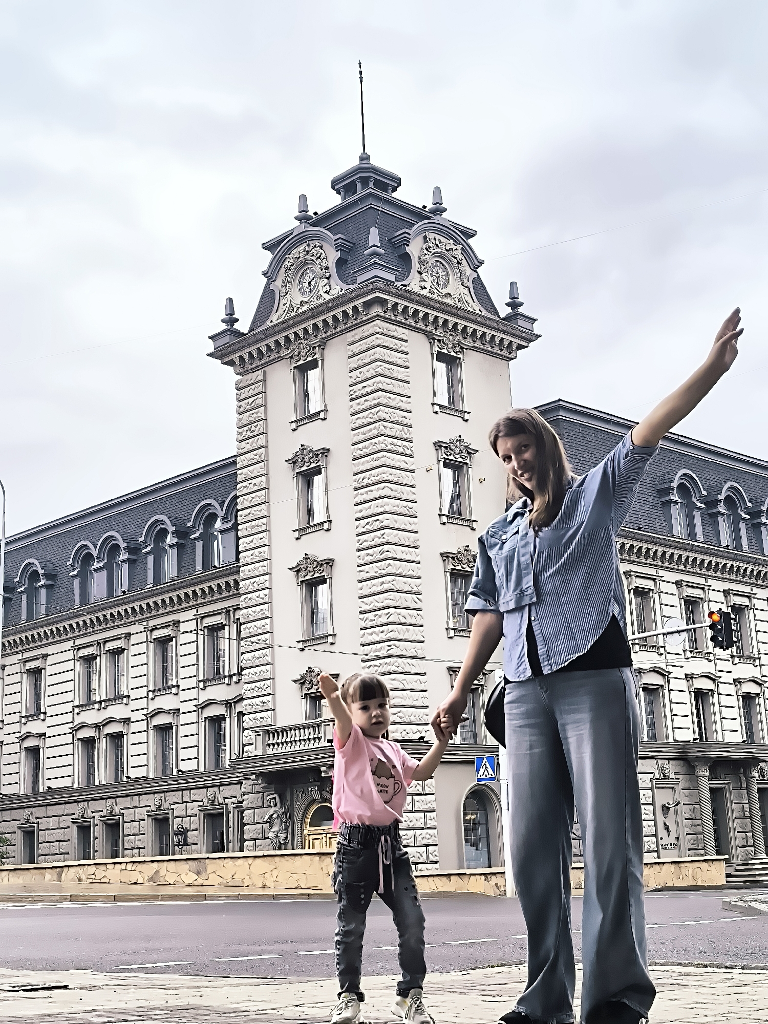 A woman and a young girl are standing on a sidewalk in front of the city administration building in Qarağandı, Kazakhstan. The building is ornate, with a mansard roof and a clock tower. The woman is wearing a denim jacket and blue striped shirt, and the girl is wearing a pink t-shirt and jeans. They are holding hands, and both appear to be posing for the camera, with their arms raised. The sky is overcast.