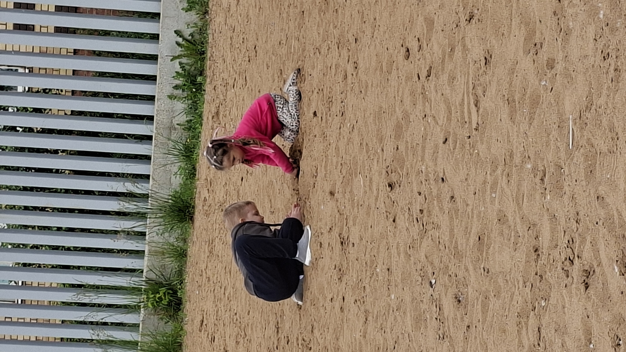 Two children, a boy and a girl, are playing in the sand near a fence in Nytva, Russia. The boy, wearing a dark hoodie and black pants, is crouched on the sand. The girl, in a pink long-sleeved shirt and patterned leggings, is kneeling and digging in the sand with her hands. Both children appear to be focused on their activity. The environment is an outdoor sandy area with a light-colored fence in the background. Green grass and some vegetation are visible behind the fence. The sand is a light brown color and appears to be somewhat damp in places. The overall scene depicts a casual outdoor activity of children playing.