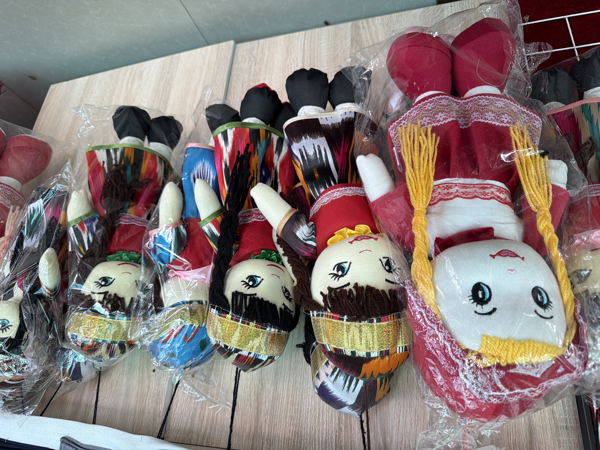 The image shows a display of several handmade cloth dolls, each individually wrapped in clear plastic and hanging from hooks against a light-colored wooden background. The dolls appear to be of various sizes and are dressed in colorful, patterned clothing that features geometric designs. Many of the dolls wear crowns or headbands with gold trim. The doll in the foreground on the left is larger than the others and has bright yellow yarn hair styled in pigtails, a red dress with white trim, and red shoes. The other dolls exhibit similar craftsmanship, with embroidered facial features and yarn hair in shades of brown and black. The environment suggests a market or shop setting, possibly in Zangiata, Uzbekistan, given the traditional attire of the dolls. There are no people visible in the frame.