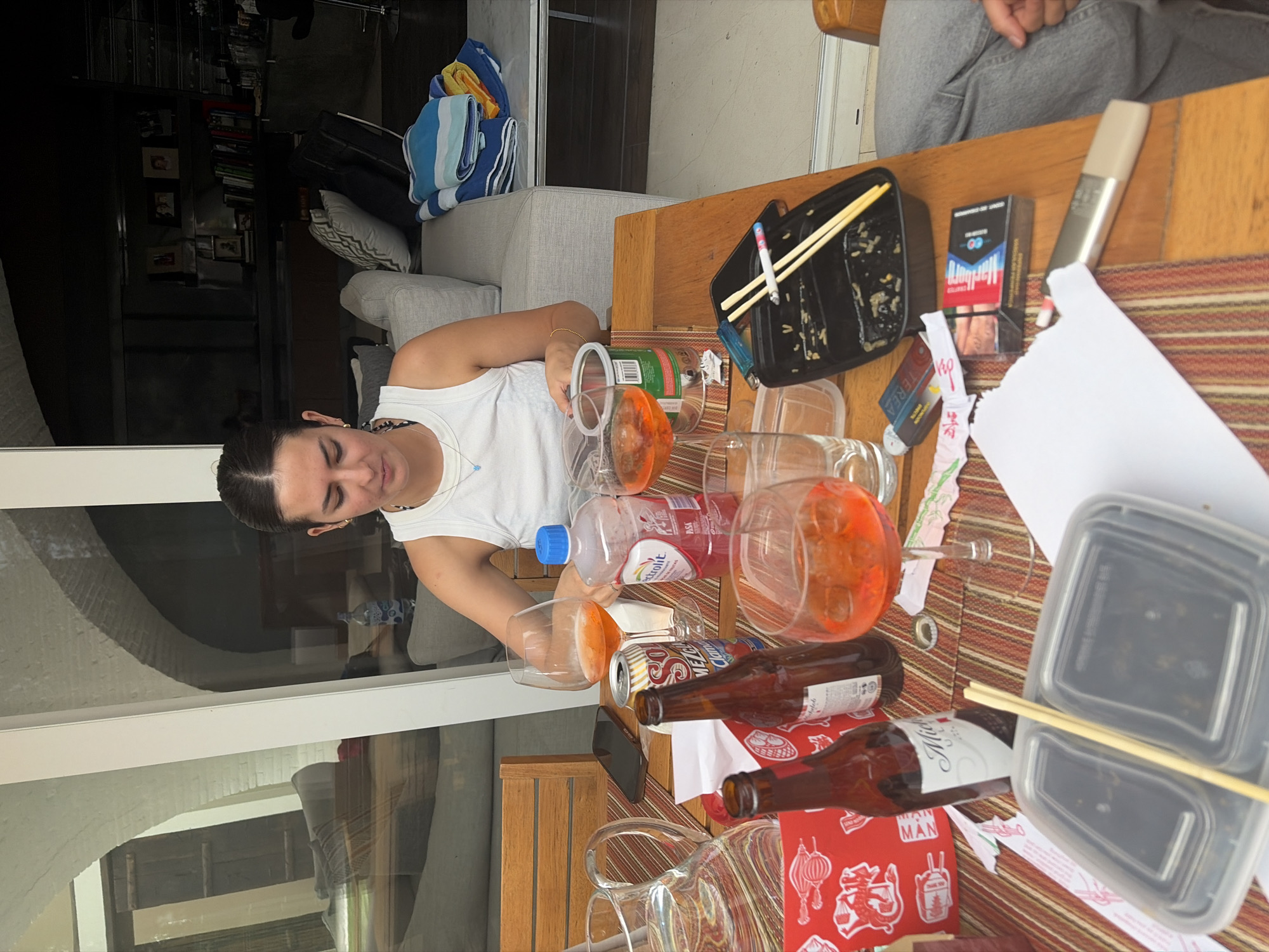 A young adult woman in a white tank top sits at a table outdoors. The table is covered with a striped runner and various items including open containers of food, empty beer bottles, drinking glasses, a juice bottle, and a pack of Marlboro cigarettes. Chopsticks are present, suggesting a meal of Asian cuisine. The location, San Bernardino Tlaxcalancingo, Mexico, is not discernable from the image. The environment visible through the glass windows suggests a residential setting.