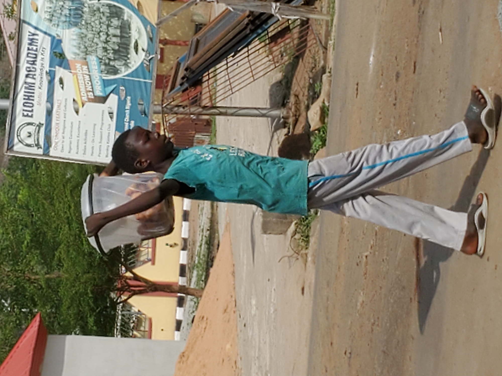 A vertical, eye-level outdoor shot depicts a young person, likely a child or teenager, walking or standing on a dirt path in Mararaba, Nigeria, during the daytime. The individual, dressed in a teal t-shirt and grey track pants with a blue stripe, is holding a large transparent container, possibly a bucket or jar, in their left hand. The container appears to hold items, though they are indistinct. The person is wearing white flip-flops and looks upward and to the right.
In the background, a large, blue, oval-shaped signboard for "ELOHIM ACADEMY" is prominent, featuring a group photo of students and text. Visible details on the sign include the motto "Knowledge is Key" and "ADMISSION IS NOW IN PROGRESS." Other partial text includes "ONE UNIQUE FEATURES" and "Nigeria." Behind the signboard, there are glimpses of green foliage, a building with a yellow facade, and a metal structure, possibly a gate or fence. The ground is a mix of light brown dirt and sand, showing some texture and small shadows. The overall lighting suggests a bright, sunny day.