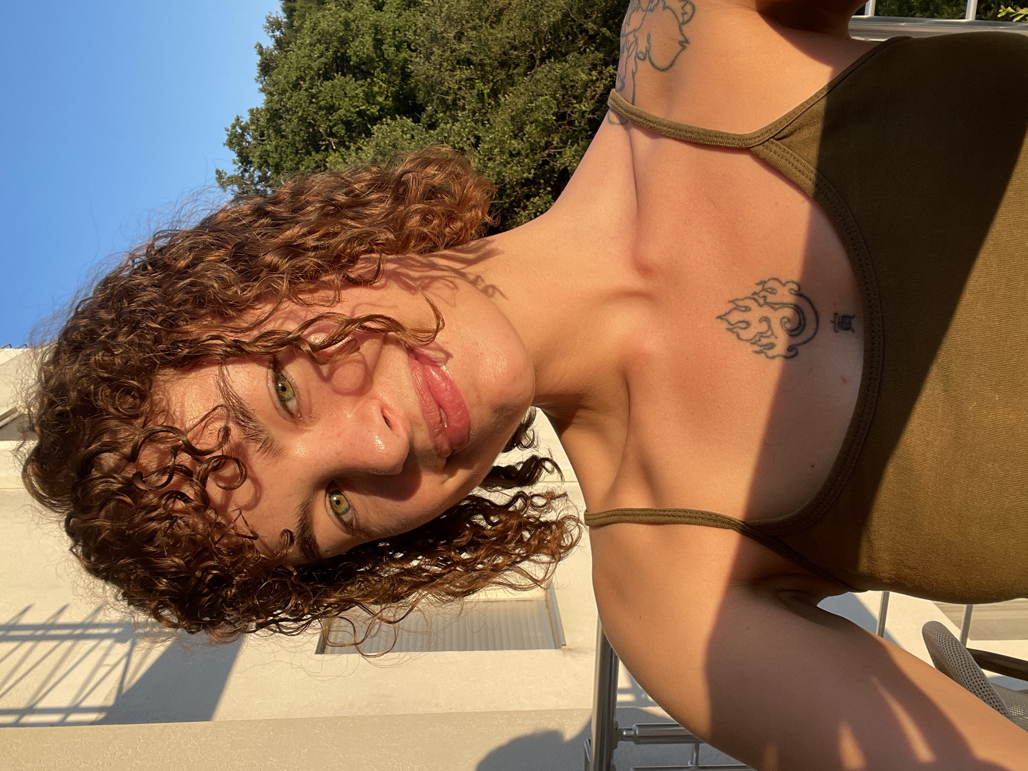 A young woman with curly, reddish-brown hair and green eyes is taking a selfie on a balcony. She is wearing a dark olive green tank top. A tattoo of flames and a crescent moon is visible on her chest, and another tattoo is partially visible on her left shoulder. The sun is low in the sky, casting a warm glow and long shadows on the wall behind her and on her face. Lush green trees and a clear blue sky are visible in the background. The balcony railing is also visible. This scene is likely in the city of Rafailovići, Montenegro.