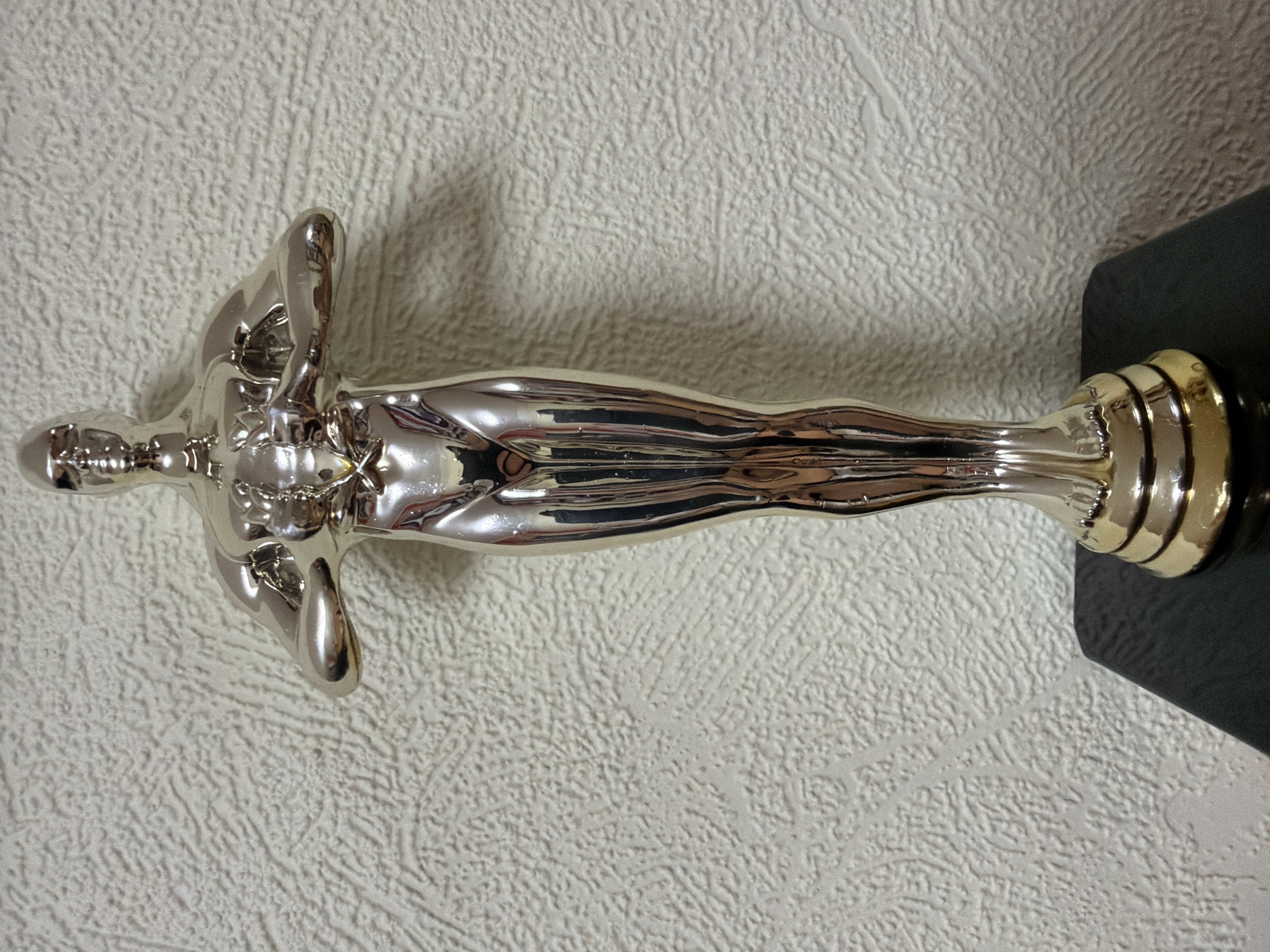 A close-up, eye-level shot shows a metallic golden statuette, resembling the Oscar award, standing upright on a black, textured surface. The statuette is positioned slightly off-center to the left, facing forward, with its arms clasped across its chest. The background is a textured off-white wall. There are no people present in the image, and no identifiable location in Kyiv, Ukraine is discernible. The lighting is even, with reflections visible on the polished surface of the statuette.