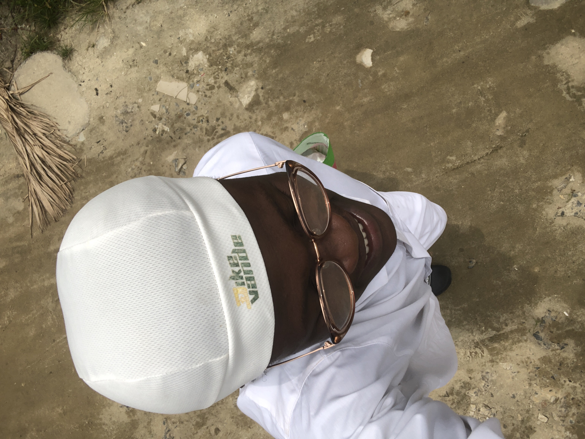 A person, viewed from above, stands on a paved surface. They are wearing a white cap with a logo that appears to say "nike vande" in green and yellow lettering. They also wear glasses with rose gold frames and a white garment, possibly a thawb. The ground is a mix of concrete and dirt, with some scattered debris and dried palm fronds in the background. The scene suggests an outdoor setting, potentially in a place like Ado, Nigeria, given the environmental cues.