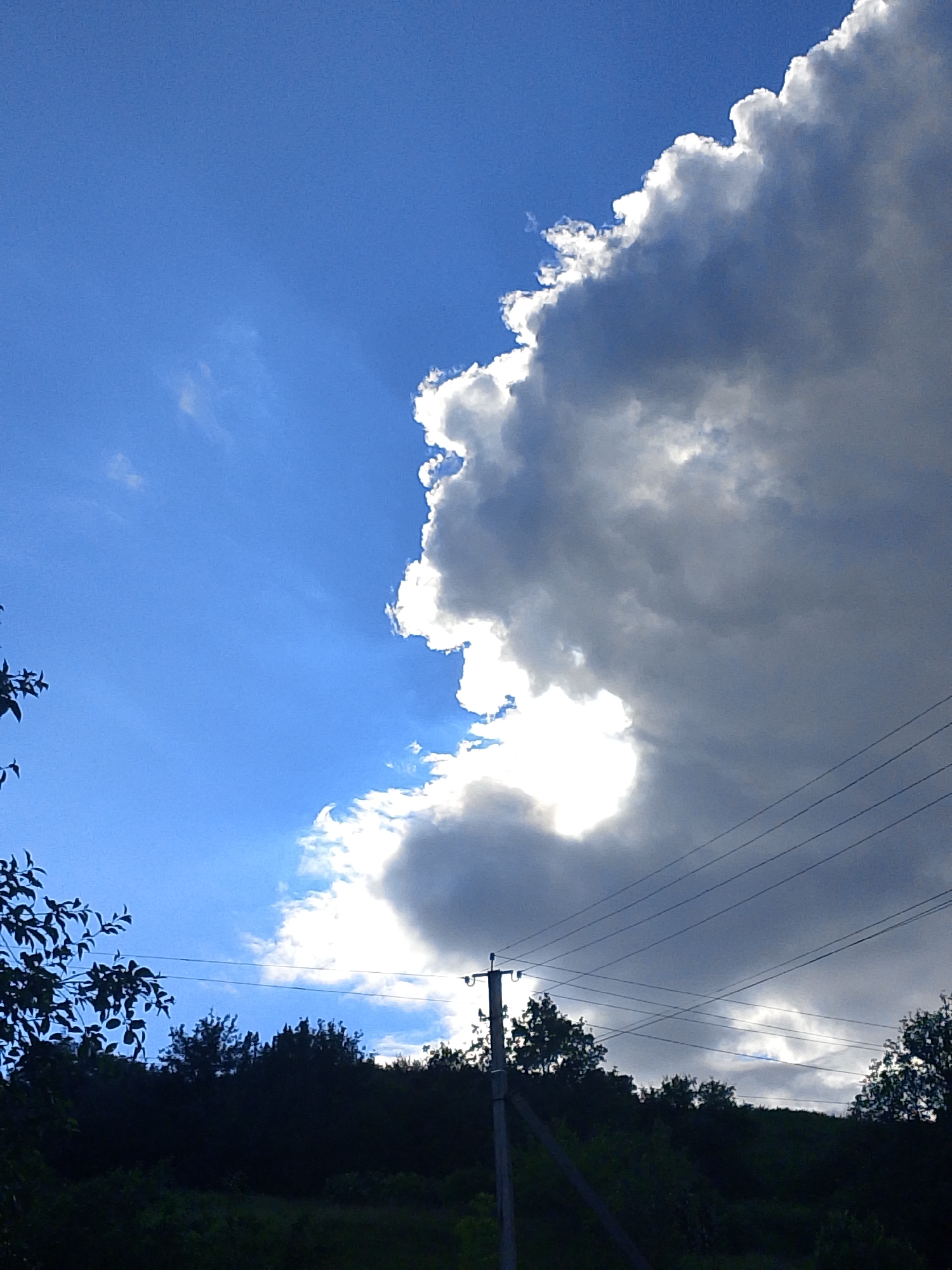 The image shows a partly cloudy sky over a landscape with trees and an electrical pole. The sky is a mix of bright blue and large, billowing clouds that are partially lit by the sun. In the foreground, there is a silhouette of trees and a utility pole with wires stretching across the frame. The location is Unknown, Unknown.