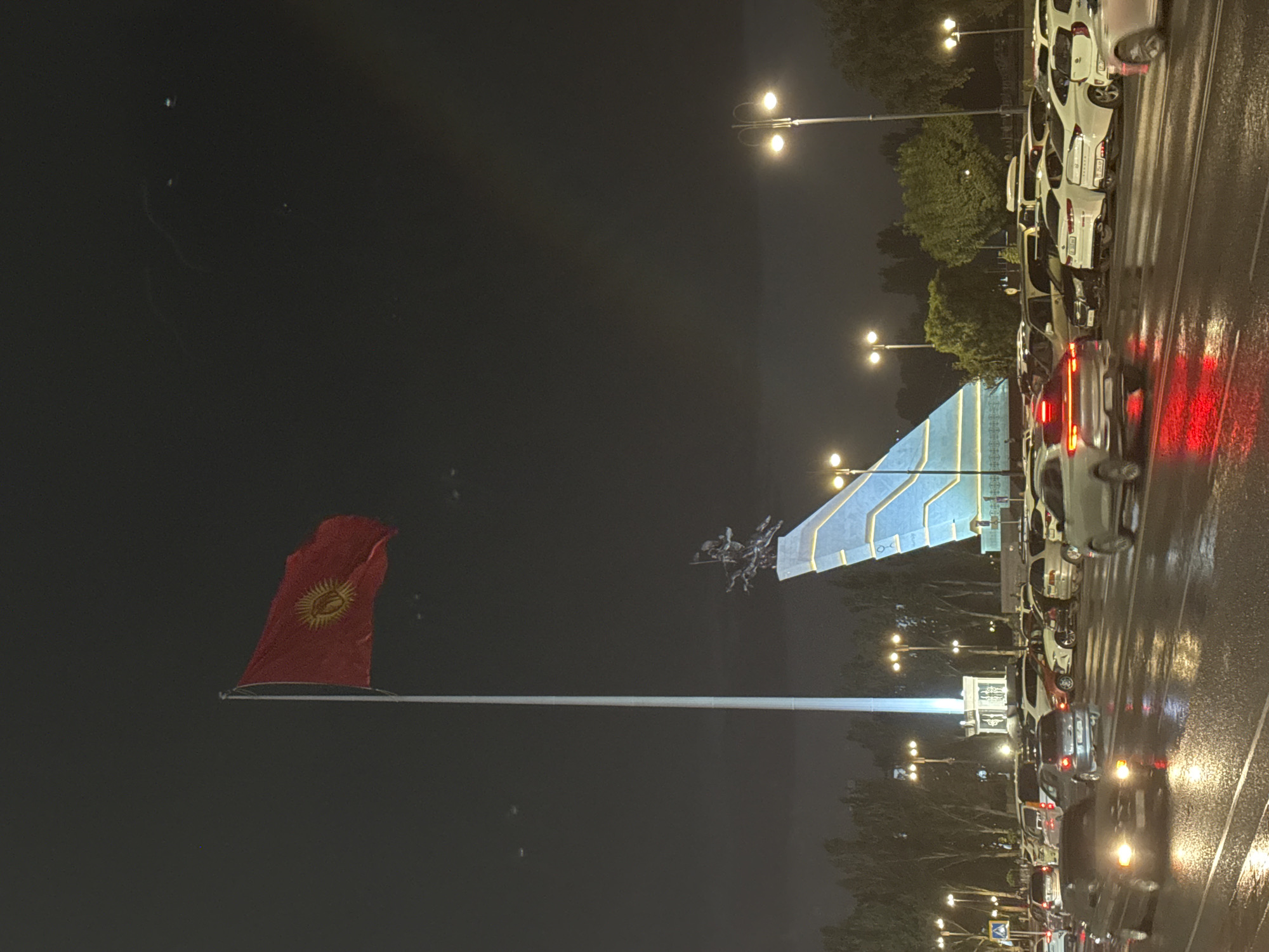 The image depicts a night scene in Bishkek, Kyrgyzstan, likely near Ala-Too Square, featuring a prominent flag pole with the Kyrgyz flag flying. A large, modern monument, possibly the "Manas" monument, dominates the background, characterized by a stylized, angled structure with blue illumination and a sculptural element of figures on horseback at its apex. Streetlights with multiple globes illuminate the area, casting a warm glow on the wet asphalt of a busy road in the foreground. Numerous vehicles, including cars and vans, are parked and in motion on the road, their headlights and taillights creating streaks of light due to the long exposure and wet conditions. Trees line the background, their forms silhouetted against the dark, cloudy night sky. The overall atmosphere is one of an urban environment at night, with elements of national pride and public space visible.