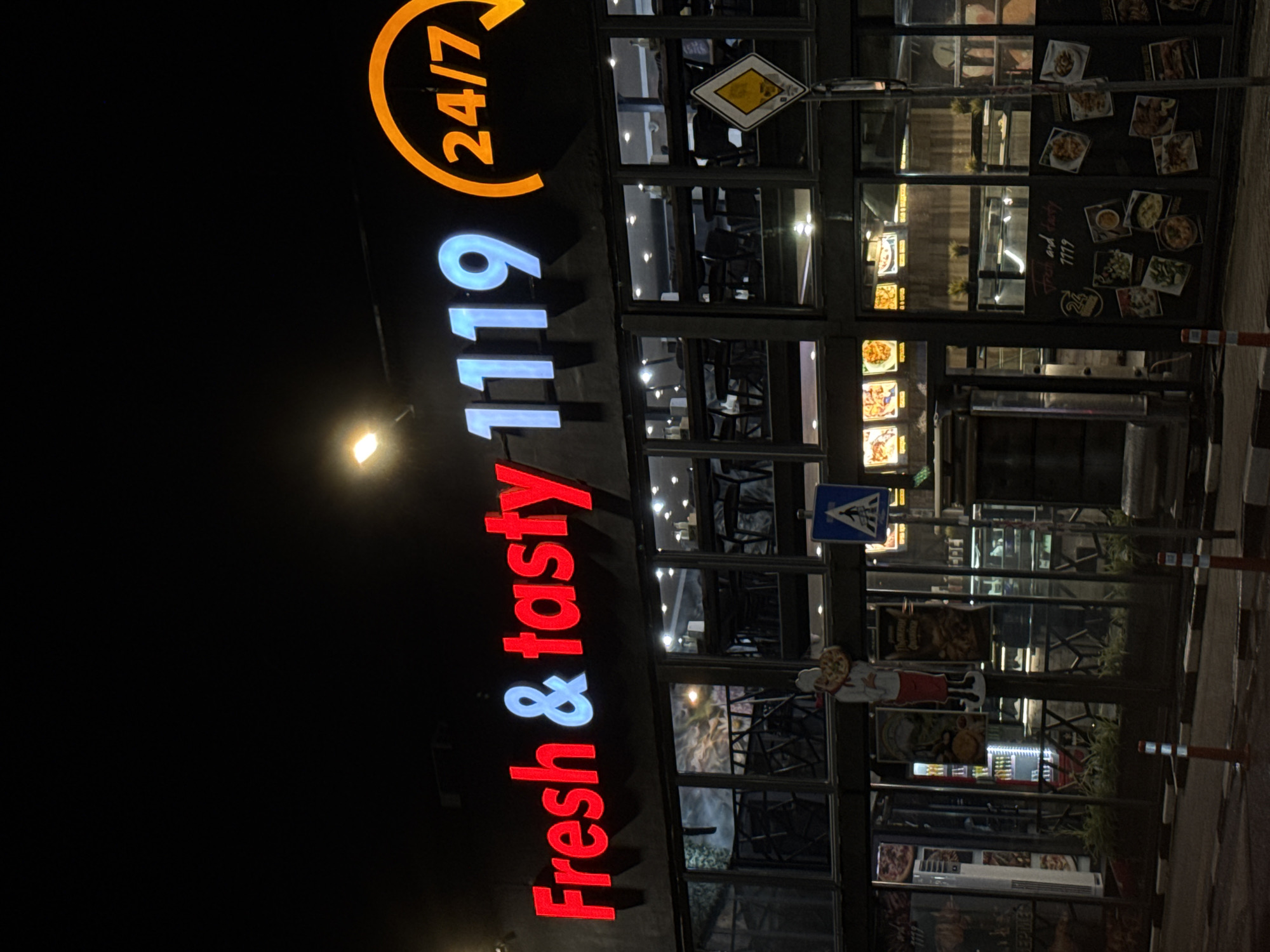Captured at night, the image depicts the storefront of "Fresh & Tasty 1119," operating 24/7. The text "Fresh & Tasty" is illuminated in red, with the ampersand in light blue, and "1119" in light blue. Above, the "24/7" logo glows in orange.
The restaurant has a large glass facade, providing a view of the interior with tables and chairs. Numerous posters showcasing food items are displayed on the windows. Street signs are also visible, including pedestrian crosswalk. The surrounding environment appears dark, indicative of the nighttime setting. The location, given the signs, is likely in Sofia, Bulgaria. There is no readily available specific location information within the image. The scene seems quiet, with no evident activity around the storefront other than the signs.