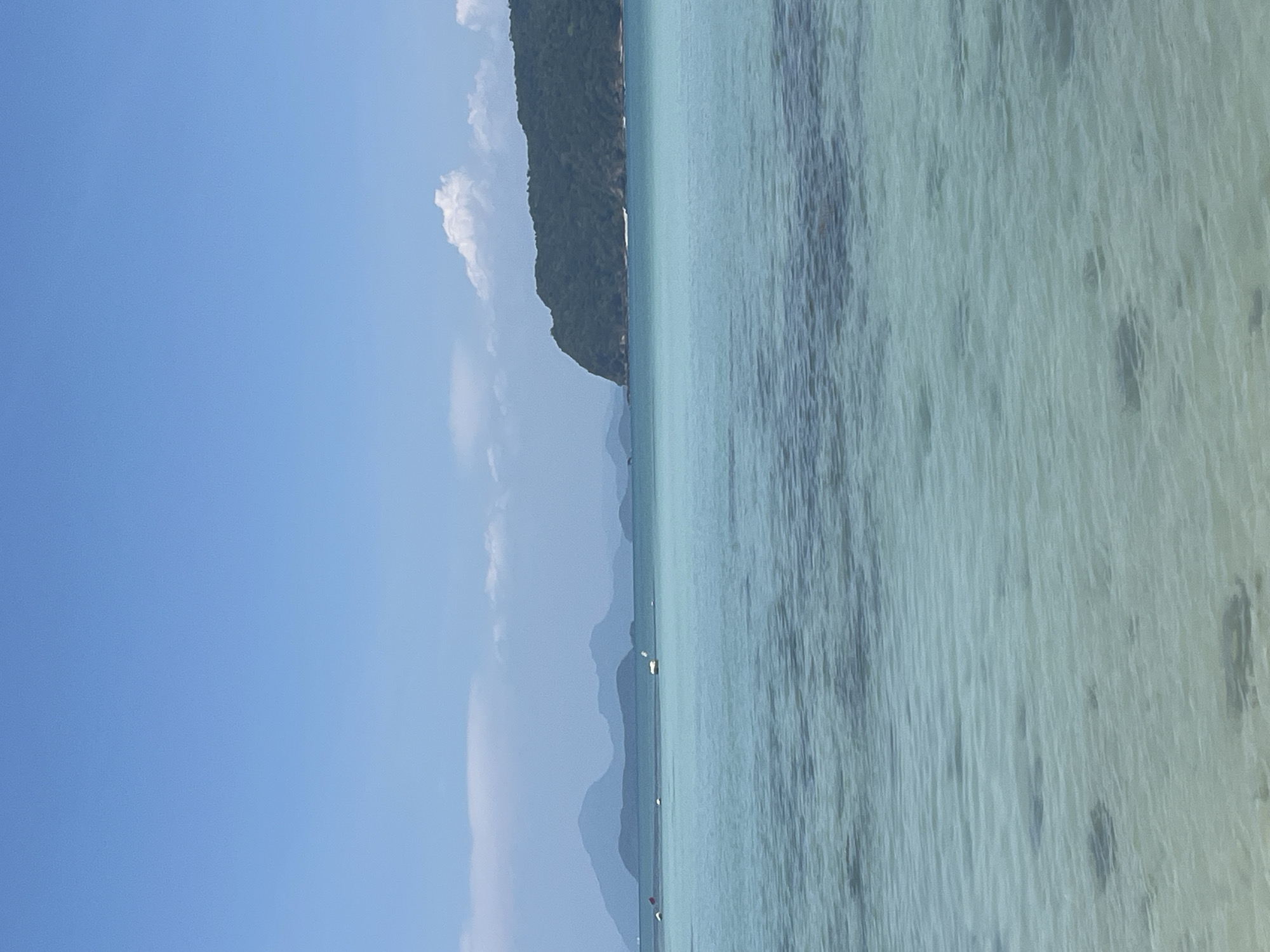 The image depicts a coastal scene in Ban Bang Khao, Thailand. The right side of the frame is filled with turquoise, clear water. The seabed is faintly visible through the water. On the left, a clear blue sky dominates, punctuated by sparse white clouds. An island covered in dense, green vegetation occupies the middle ground. Faint silhouettes of other islands are visible in the distance.