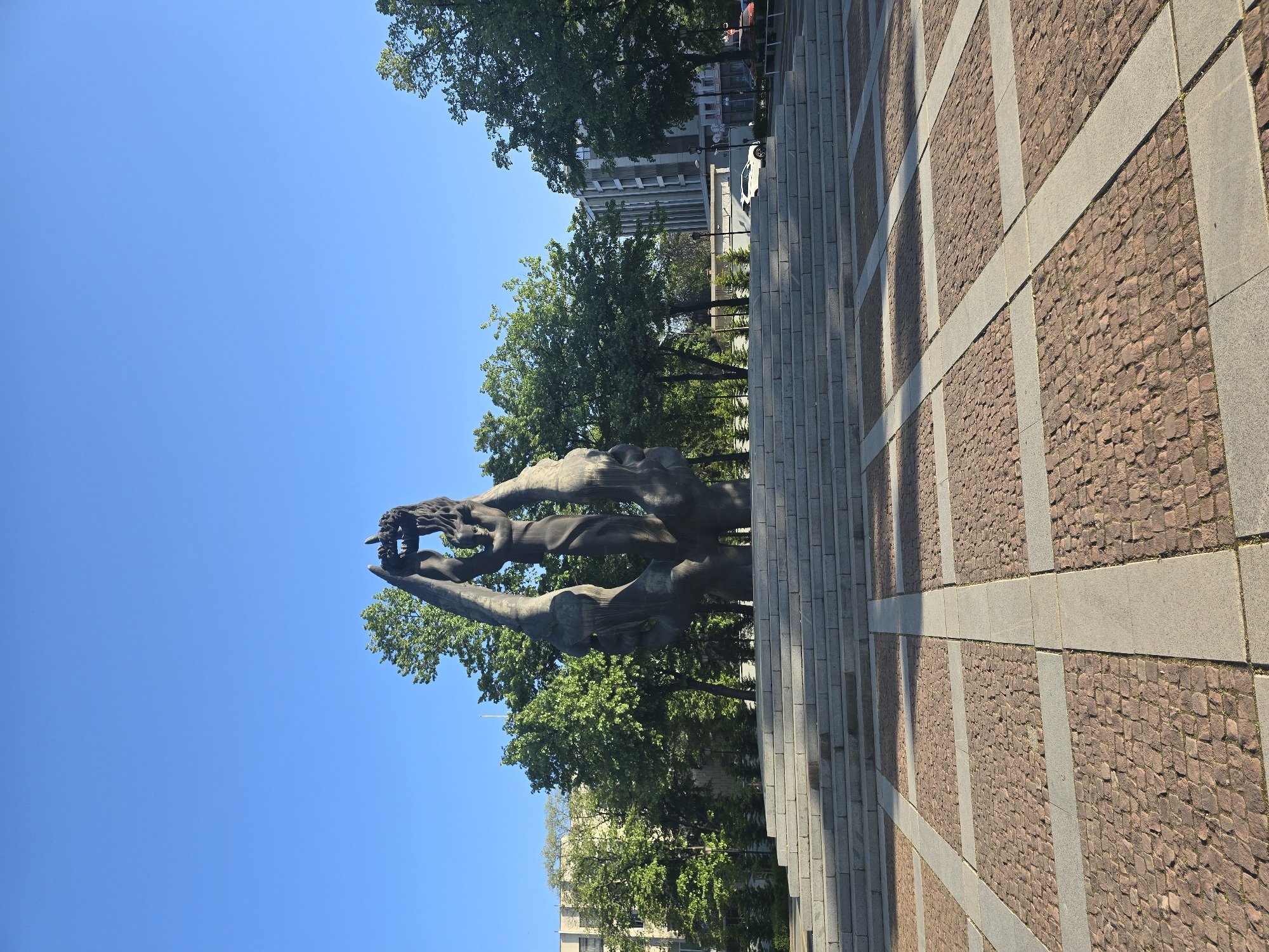 The image displays an outdoor scene featuring a large, dark-colored, abstract sculpture under a clear blue sky. The sculpture is monumental, with dynamic, elongated forms that appear to be reaching upwards, possibly representing figures or mythical creatures. It is made of a dark material, likely metal or stone, and stands amidst green foliage from numerous trees.
Behind the trees in the mid-ground, multi-story buildings of a modern architectural style are visible, suggesting an urban setting. A white car is partially discernible on a street or parking area near these buildings.
In the foreground and extending to the right, there is a patterned paved surface. This pavement is composed of wide, light-grey rectangular slabs that form a grid, with the spaces within the grid filled with smaller, reddish-brown cobblestones or pavers. Distinct, long shadows are cast across this paved area, indicating strong sunlight.
The sky is a vibrant, unblemished blue, signifying clear weather. The presence of strong shadows suggests it is daytime, likely mid-morning or early afternoon. No people or specific activities are visible, presenting a static and serene view of the public space. No text is visible in the image.