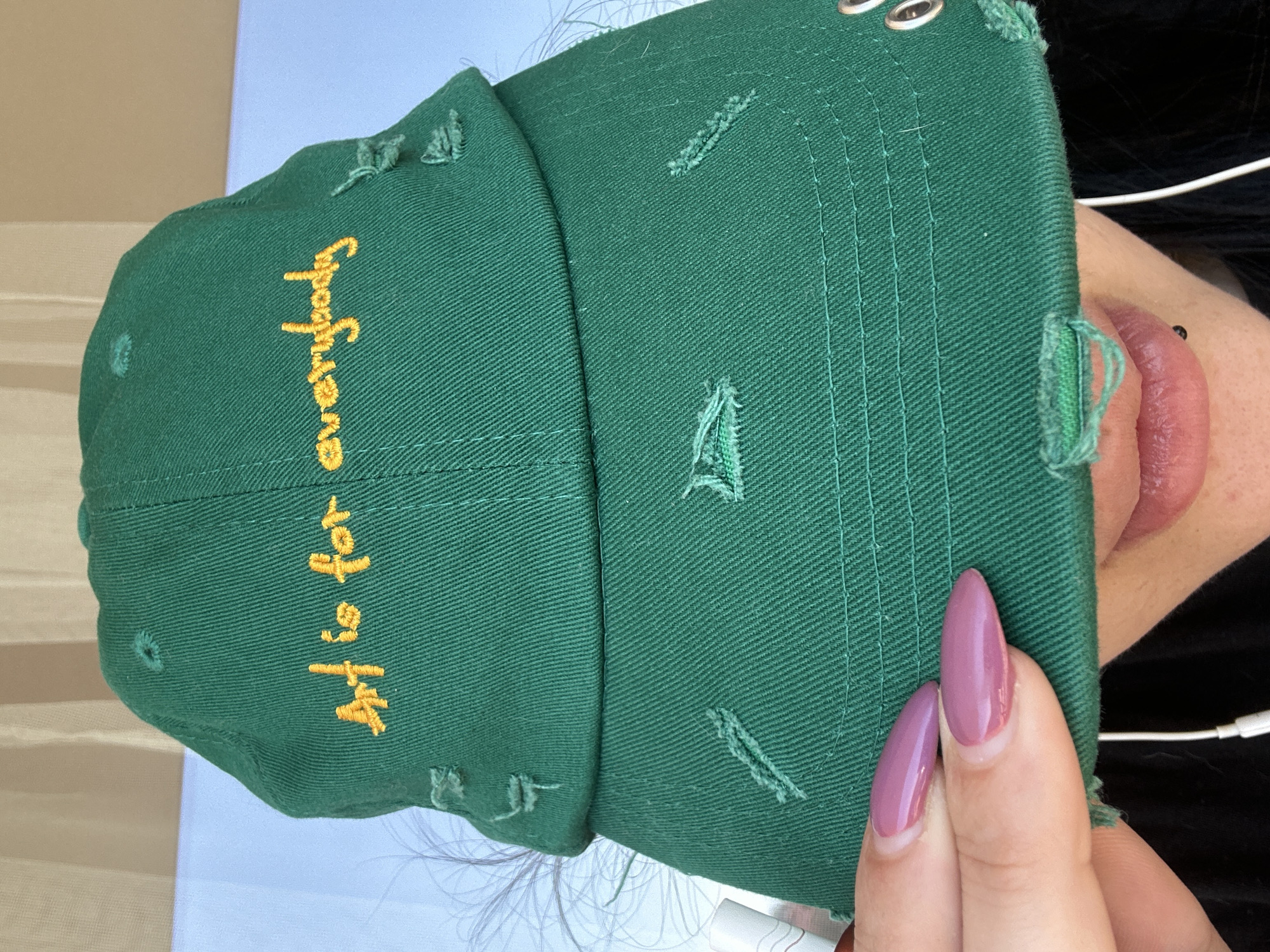 The image depicts a person holding a distressed green baseball cap with the text "Art is for everybody" embroidered in yellow. The cap is close to the person's face, with a portion of their lip and a visible lip piercing. Their hand is visible, displaying elongated pink nails. The background consists of a pale blue surface, possibly a table or counter. The location is not identifiable as Varna, Bulgaria, from the provided image.