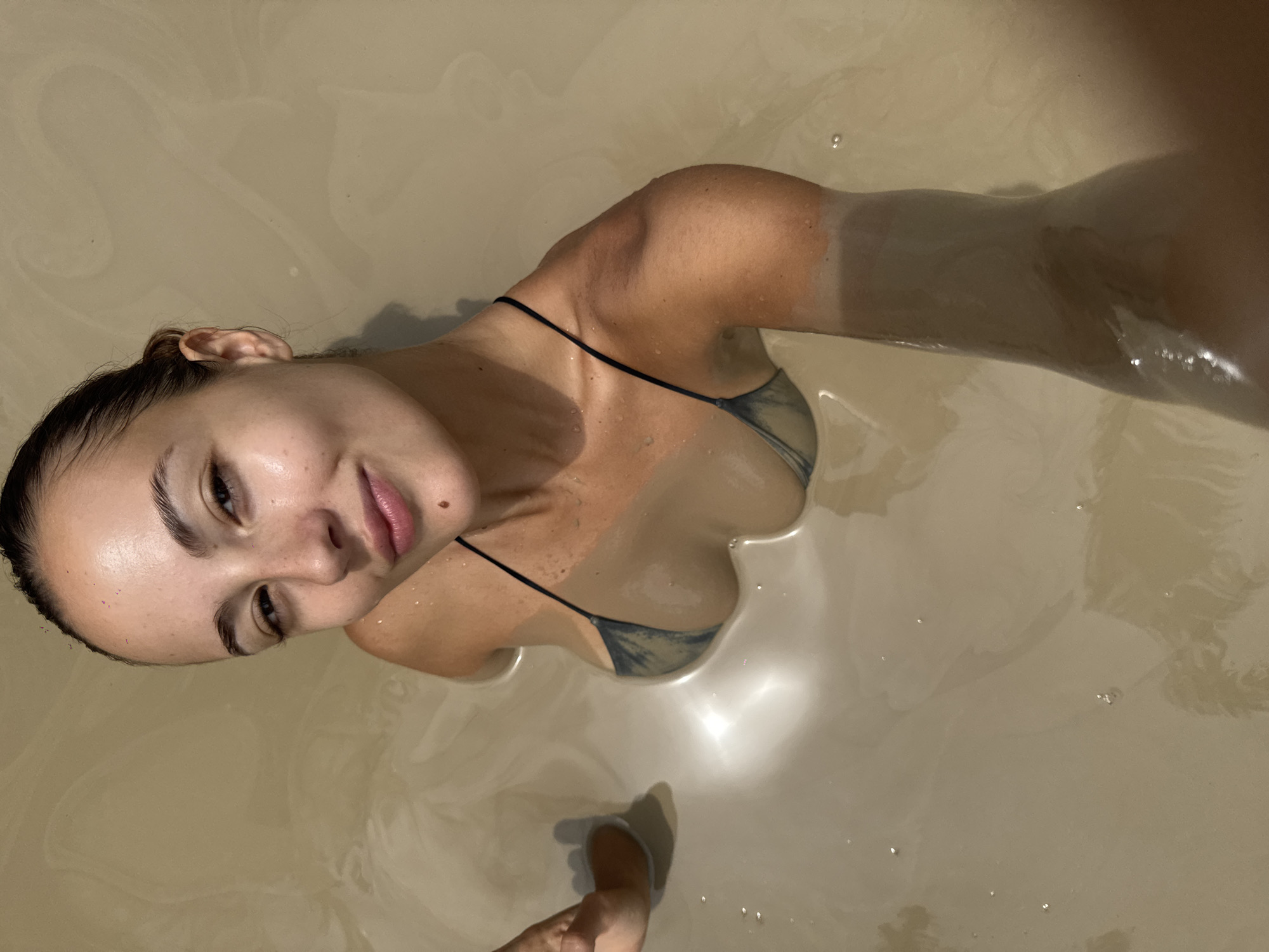 A woman is photographed from above in what appears to be a muddy bath. She is wearing a two-piece swimsuit. The location may be in Ban Khlong Khian, Thailand, but this cannot be confirmed from the image itself. Her body is partially submerged in the opaque, brown liquid. A foot is visible in the lower portion of the image.