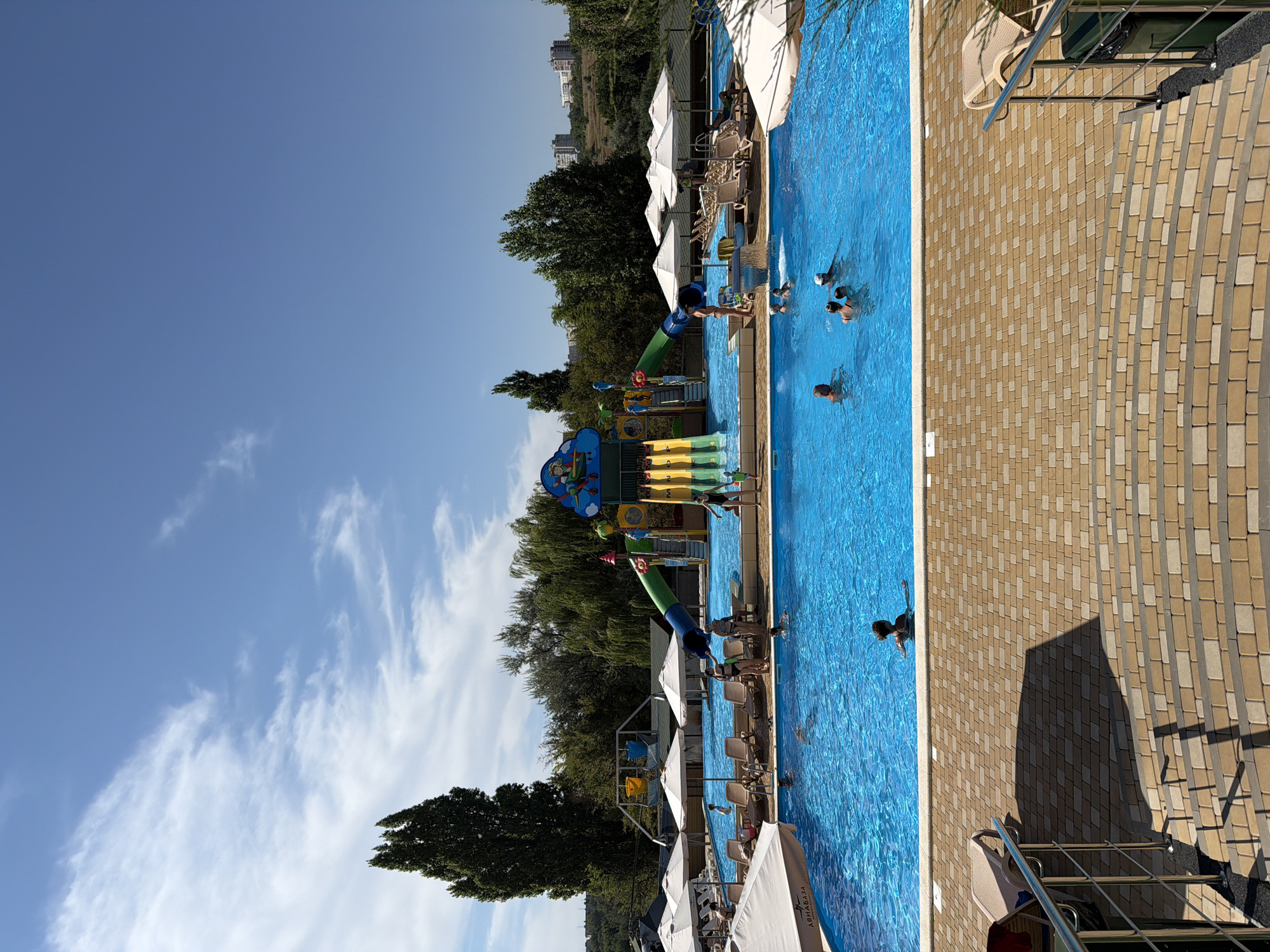 The image depicts a vibrant outdoor water park on a sunny day, likely in Kamyshin, Russia, given the inscription "АВИАБАЗА" (Aviabaza) visible on a large white umbrella in the foreground, which is the name of a local water park in Kamyshin.
In the center of the frame, a large, bright blue swimming pool dominates the scene, with several people enjoying the water. Children and adults are visible swimming and wading. To the left of the pool, a series of white umbrellas provide shade over lounge chairs, some of which are occupied.
At the far end of the pool, a colorful water slide complex is the main attraction. It features multiple slides, including a large green tube slide and yellow slides with cascading water. Several figures are seen interacting with the slide structure, either climbing or preparing to slide.
The environment is lush and green, with tall trees lining the perimeter of the water park. In the background, rolling hills with buildings suggest the park is situated on the outskirts of a town or city. The sky is a clear, bright blue with scattered white clouds, indicating pleasant weather conditions.
The foreground shows a textured, paved walkway with curved steps leading down to the pool area. The paving stones are a mix of beige and brown, creating a patterned effect. Metal railings are present along the steps and the edge of the walkway.