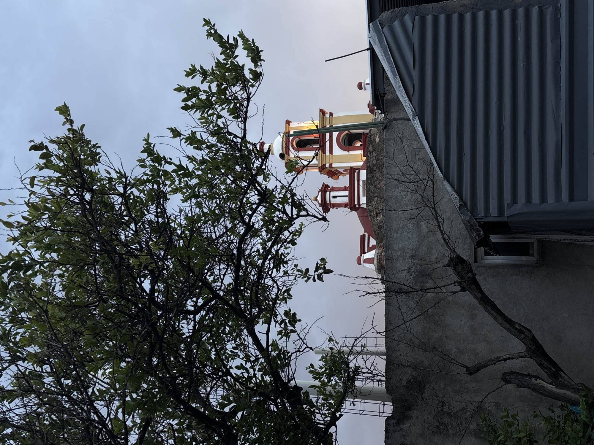This image depicts a static urban scene in San Andrés Cholula, Mexico, under low light conditions consistent with dusk or dawn. The central feature is the upper portion of a church, specifically a bell tower and a smaller domed structure, identified as part of the Parroquia de San Andrés Apóstol. The main bell tower is predominantly white and yellow with red architectural details, featuring arched openings where an internal light source is active. A smaller, red and white domed structure is positioned to the left of the main tower.
In the foreground, the upper left quadrant is dominated by the dense, green foliage and dark branches of a deciduous tree. A rough, textured grey concrete wall extends horizontally across the midground, partially obscuring the lower sections of the church and adjacent structures. A bare, dark-barked tree with multiple branches extends upwards from the lower left, positioned in front of this wall. To the lower right, a structure with a steeply angled dark grey corrugated metal roof is visible, covering part of a building. A small, dark rectangular window with a light frame is set into a wall section beneath this roofline. Behind the concrete wall and to the left of the church structures, white cylindrical poles and dark metal scaffolding are partially visible. The sky in the background is a uniform light blue-grey, indicating an overcast or transitional lighting period. No people or animals are present in the scene.