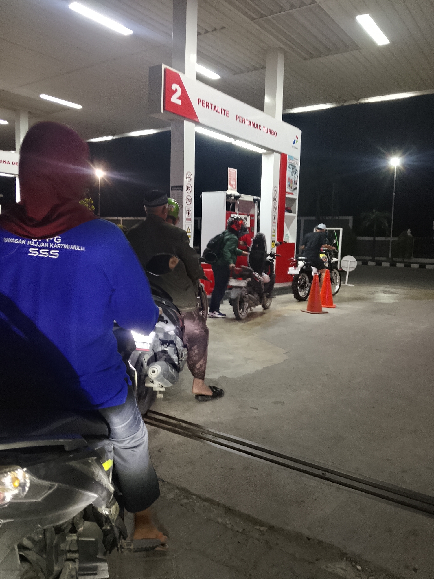 This image depicts a nighttime scene at a Pertamina gas station, likely located in Kasiman, Indonesia, given the textual cues on the signage. The central focus is a red and white gas pump island. In the foreground, the back of a person wearing a blue long-sleeved shirt with white text and a red hood is visible, seated on a motorcycle. Further back, near the gas pumps, are several other individuals on motorcycles. One person, wearing a green helmet and jacket, is standing by a motorcycle. Another individual in a dark jacket and patterned pants is seated on a motorcycle. Two orange traffic cones are placed on the ground near a white motorcycle. The lighting suggests it is after dark, with overhead fluorescent lights illuminating the gas station canopy. The ground appears to be concrete pavement.