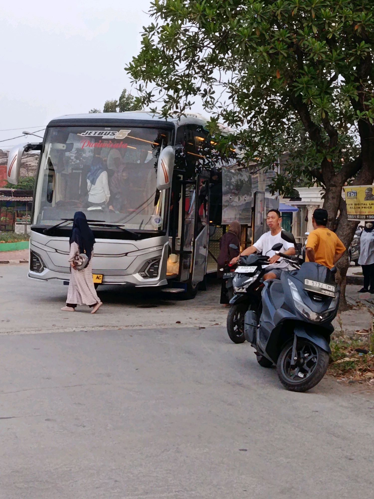 A wide shot captures a street scene in Bekasi, Indonesia, with a large passenger bus and several motorcycles as the main subjects. The bus, identifiable by "JETBUS" branding, is silver and has its doors open, suggesting it is either boarding or disembarking passengers. A woman in a long, patterned dress and a dark hijab walks away from the bus towards the foreground. Inside the bus, other passengers are visible through the windows.
In the middle ground, two men are on motorcycles. One man, wearing a white t-shirt and shorts, is seated on a dark-colored scooter, looking towards his right. Another man, in an orange t-shirt and a black cap, stands beside him with his hands on his hips. A third, darker-colored scooter is parked in front of them.
A tree with lush green leaves is prominent on the right side of the frame, partially obscuring a sign with some text and images. The background reveals buildings and a cloudy sky, typical of an urban environment. The ground is paved, suggesting a road or a parking area. The overall atmosphere appears to be casual, possibly a transit point or a roadside stop.