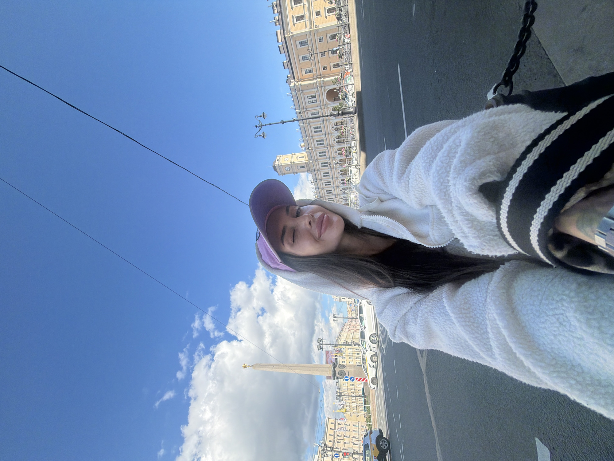 A woman is taking a selfie outdoors on a sunny day in Saint Petersburg, Russia. She is wearing a white sherpa hooded jacket and a purple baseball cap. She is winking and smiling at the camera. In the background, there is a large obelisk with a star on top, a building with a clock tower, and other city buildings. Cars and a street light are also visible. The sky is blue with some white clouds. The woman's left hand is visible, holding a black strap with white stripes. There is a chain in the foreground to the right.