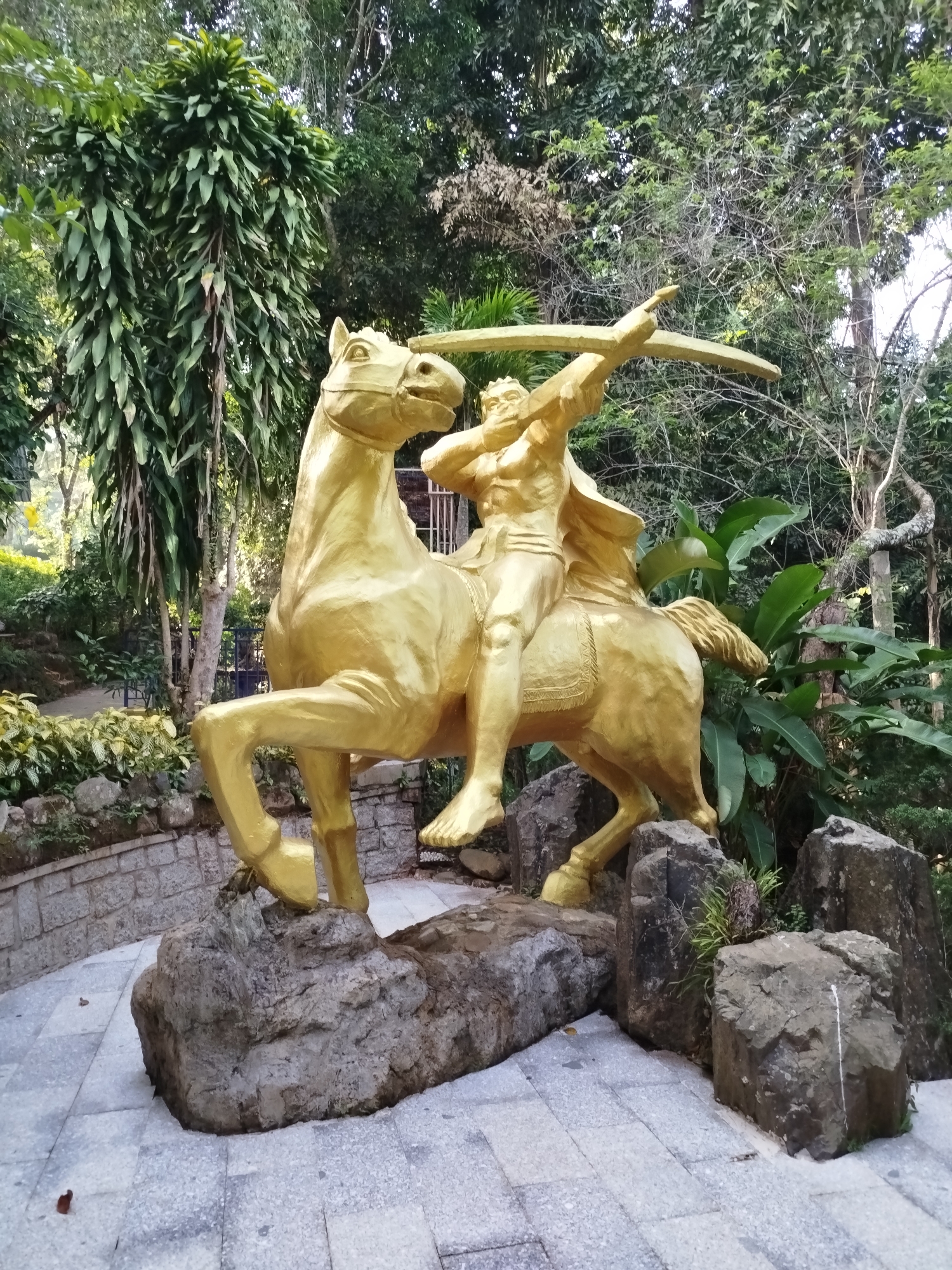 A golden statue of a bare-chested man on horseback, drawing back a bow and arrow, stands on a paved area surrounded by rocks and lush greenery. The statue is the focal point, with the man depicted as muscular and the horse in a dynamic pose. The paving stones are gray and geometrically arranged. Rocks are placed around the statue as part of the display. A variety of plants and trees form the background, suggesting a park or garden setting. The photograph depicts a statue of D'ran, a K'Ho hero in Da Lat, Vietnam.