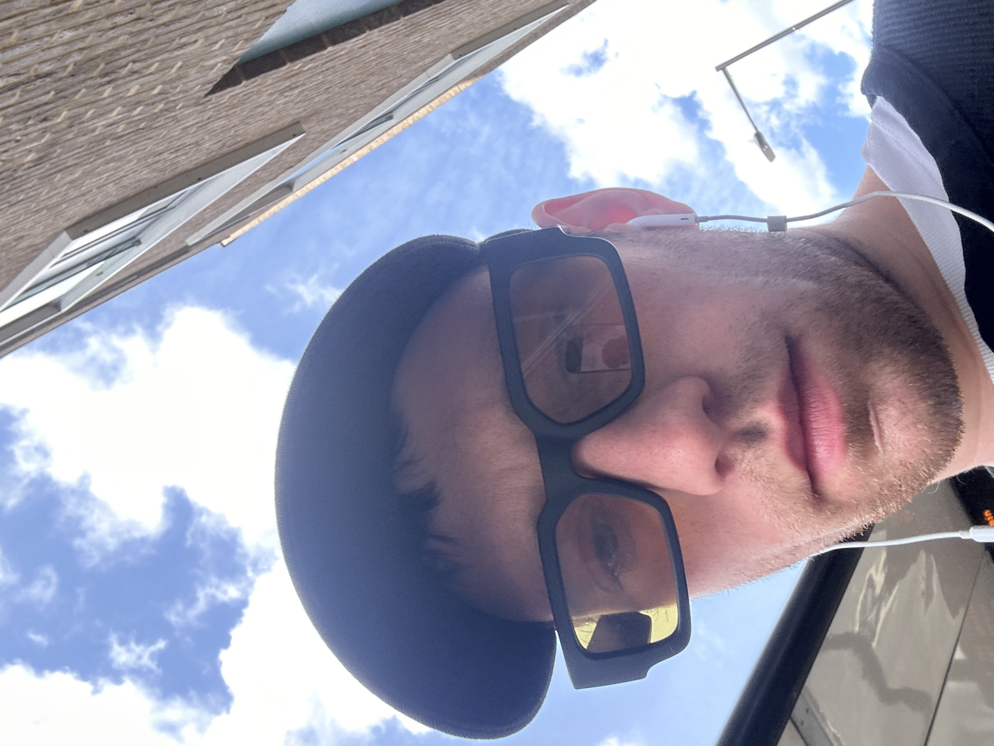 A close-up, low-angle shot shows a fair-skinned individual wearing a black hat and sunglasses with a thick black frame. The person has short brown hair, a beard and is wearing earphones. The sky is blue with scattered clouds. In the background, a section of a building with brickwork and white framed windows is visible, along with a street light. The image does not contain any information that would identify the specific location as London.