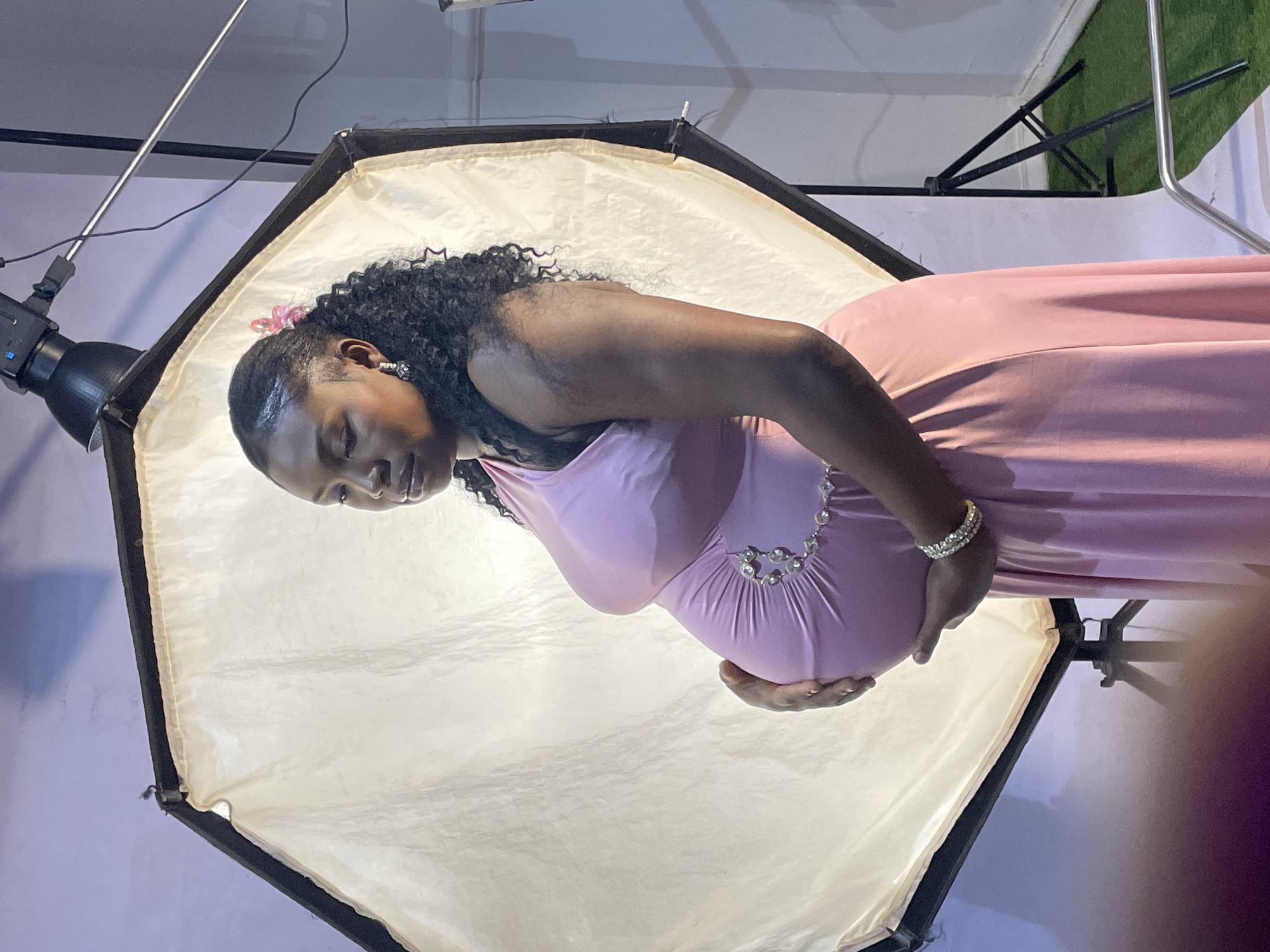 A photograph shows a pregnant Black woman in a pink dress standing in front of a softbox lighting setup. Her hands rest on her pregnant belly. Her eyes are closed. The background suggests an indoor studio environment. There are no other identifiable details to ascertain a specific location in Jos, Nigeria.
