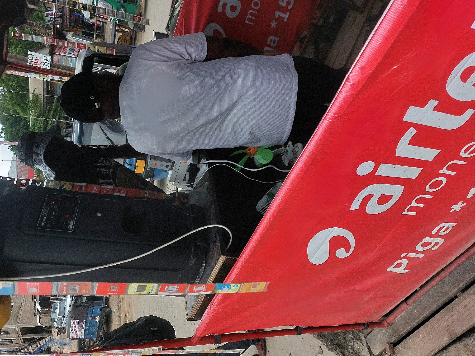 Here's a description of the image:
**Scene Overview:** The image appears to be taken at a small business location, likely a vendor's stand, in Dar es-Salaam, Tanzania. The photo is slightly rotated.
**People:** There are two men in the shot. One is wearing a white t-shirt and a black baseball cap, and is turned away from the camera, possibly interacting with something out of view. Another man, only partially visible, wears what looks like a patterned hat.
**Objects:**
* A large black speaker labeled "Skyucod".
* A red banner with white text advertising "Airtel money".
* SIM cards are visible.
* A small toy fan.
* Vehicles, including a "Bajaji" (Tuk-tuk) in the background.
* Various signs and advertisements, one sign appears to have numbers "7972".
**Actions/Interactions:** It appears the men are working at the stand, possibly involved in selling or registering SIM cards or mobile money services.
**Environment:** The scene is outdoors, likely a busy area with shops and vehicle traffic. It is daytime. The style of signs, vehicles and the advertisement point to a location in Tanzania, specifically Dar es-Salaam.