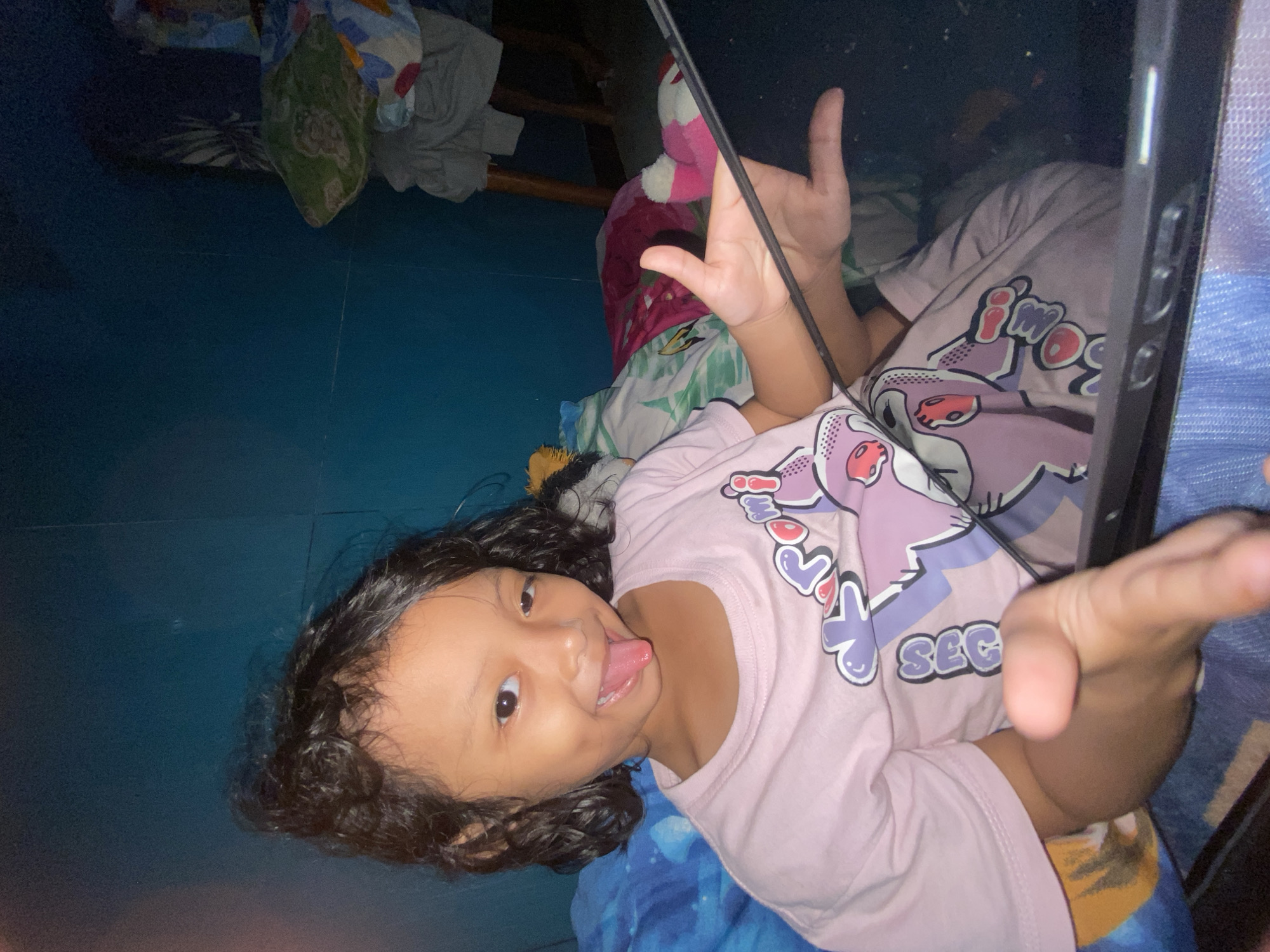 A young child with dark curly hair is lying on a blue and yellow patterned bedspread, holding a dark-colored tablet or phone. The child wears a light pink t-shirt with a cartoon design. The child is looking towards the camera and sticking out their tongue. Other items, including folded clothes, a wooden structure (possibly a bed frame), and other textiles are visible in the background. The location is possibly Gelam, Indonesia, based on prior context.