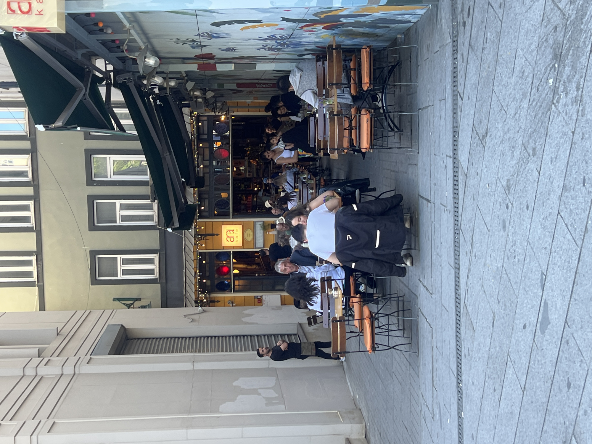 The image shows an outdoor dining area in Beyoğlu, Turkey. Diners are seated at tables along a narrow street, with buildings lining both sides. An awning shades the tables. A waiter stands near the front of the restaurant. The street is paved with stone.
