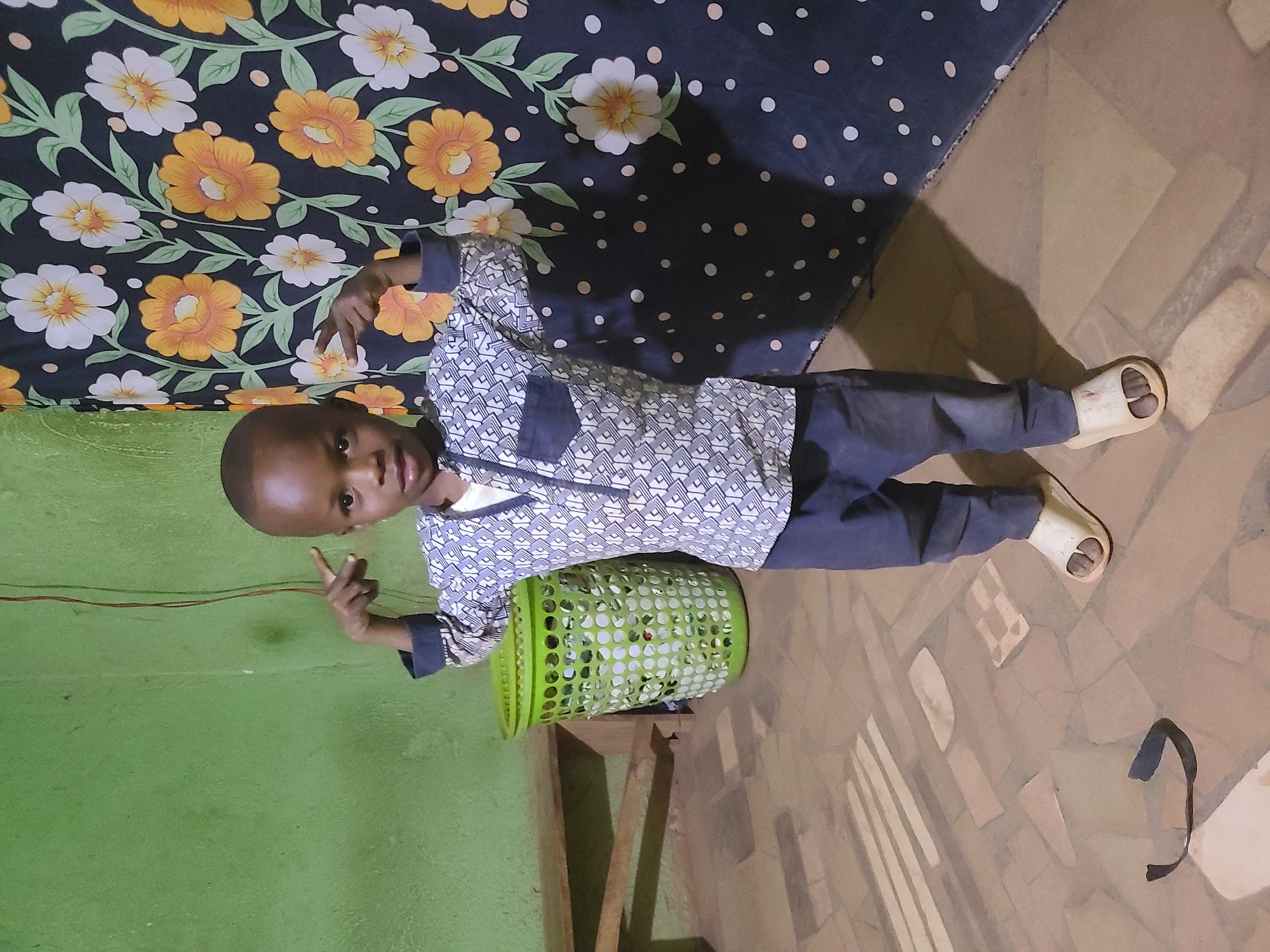 A young child stands indoors near a green wall and a floral-patterned curtain. The child is wearing a patterned shirt and dark pants, giving a peace sign with one hand. A green plastic laundry basket is positioned near the child. The floor consists of light brown tiles.
