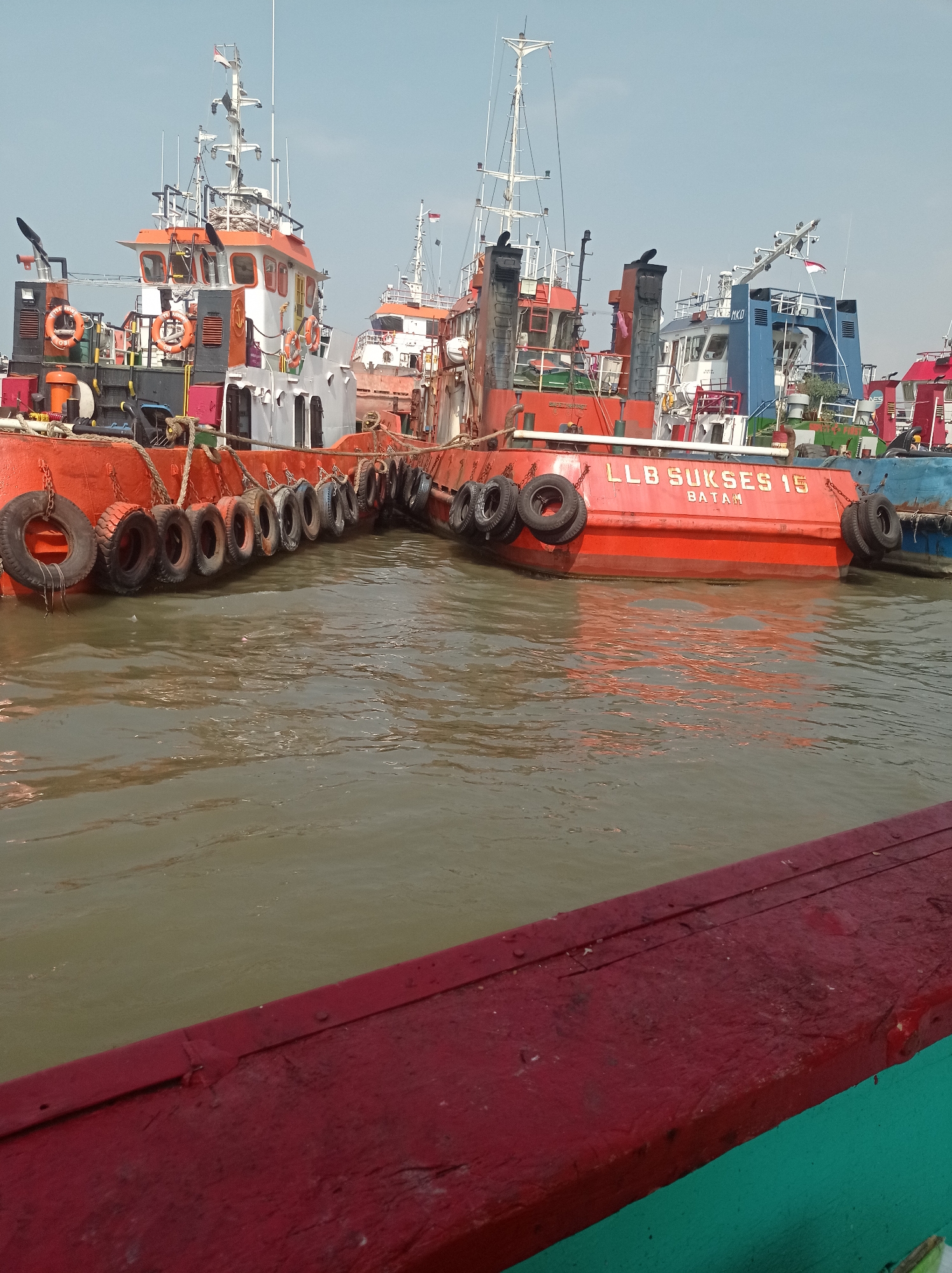 This is a medium shot, eye-level, outdoor photograph taken on a clear day. The scene depicts a harbor with several tugboats moored together. The foreground is dominated by the edge of a red wooden railing, with a sliver of teal-colored painted surface visible below it. The water is a murky green-brown color with moderate ripples, reflecting the light and the undersides of the boats.
In the midground, two large orange tugboats are prominently featured. The tugboat on the left has a white and orange superstructure with several windows. It is equipped with life preservers. The tugboat on the right is larger and more heavily laden, with an orange hull. Its superstructure is mostly orange and gray, with a prominent blue structure behind the main cabin, housing a crane or other equipment. The name "LLB SUKSES 15 BATAM" is painted in gold lettering on the hull of this tugboat. Both tugboats have numerous black tires hanging vertically along their sides as fenders.
Behind these two tugboats, other vessels can be seen. To the left, parts of another orange and white tugboat are visible. To the right, a blue and white vessel with a large blue A-frame structure is partially visible, and further back, a white vessel with a prominent mast and a red hull can be discerned. The sky is a pale blue with no visible clouds. The photograph appears to have been taken from another vessel or dock. Given the context provided, this scene is located in the city of Tegal, Indonesia, likely within its harbor.