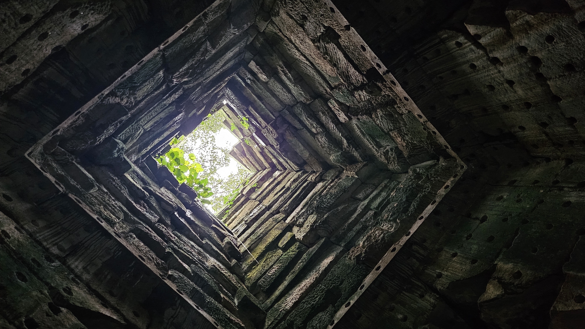 This image shows a view looking up through a square opening in the stone ruins of an ancient temple, likely Ta Prohm, in Siem Reap, Cambodia. The stone walls are constructed of large, rough-hewn blocks, stacked in layers and showing signs of age and weathering, with moss and lichen growing on them in places. The opening at the top reveals a bright, overcast sky and the green foliage of trees and vines that have grown through and around the structure, suggesting the encroachment of nature upon the ancient architecture. The perspective from below emphasizes the towering height and the angular, converging lines of the stone walls, creating a sense of depth and mystery. There are no people or animals visible in the frame.